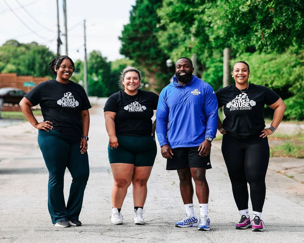 Three clients standing outdoors with Founder, Micah Baisden, smiling, with greenery in the background. The three clients are wearing black PowerHouse Sports Academy t-shirts, and Micah is wearing a blue jacket with the PowerHouse Sports Academy logo.