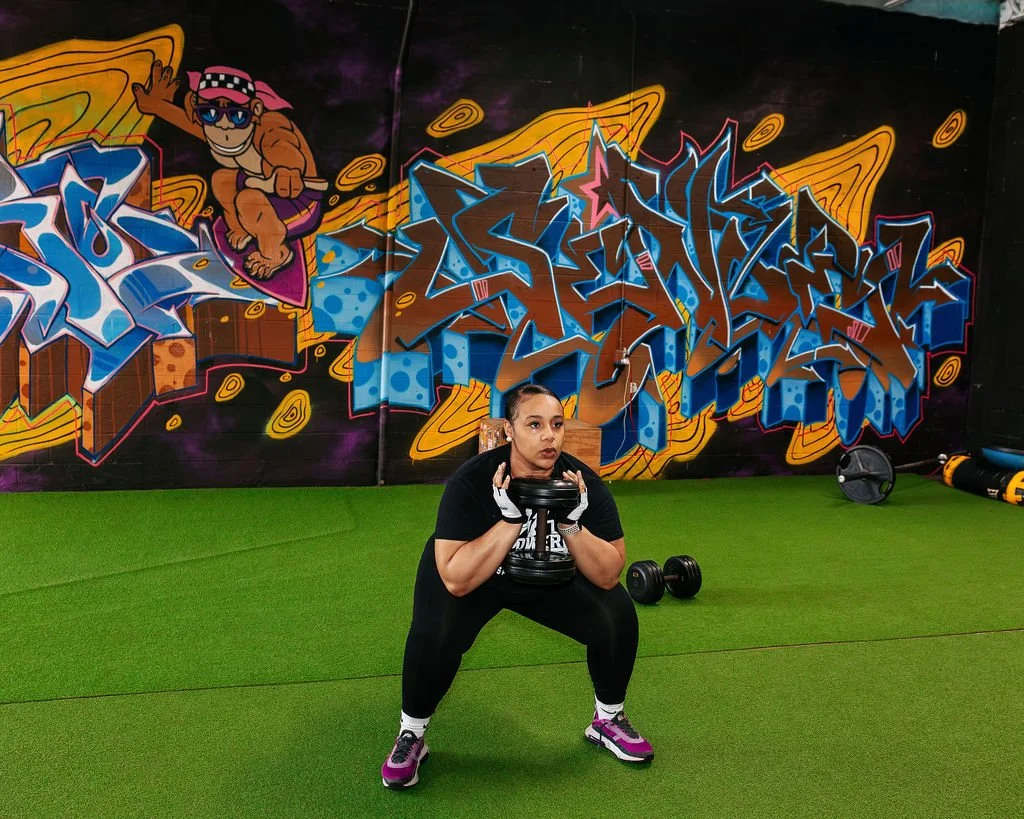 A client doing squat exercises with a weight plate in the gym. The gym has green turf flooring and a graffiti mural on the wall featuring a cartoon character and vibrant abstract designs.