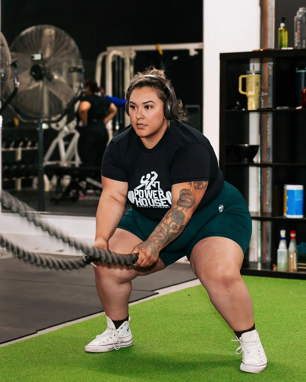 A client wearing headphones is performing a battle rope exercise in the gym. The gym has green turf.