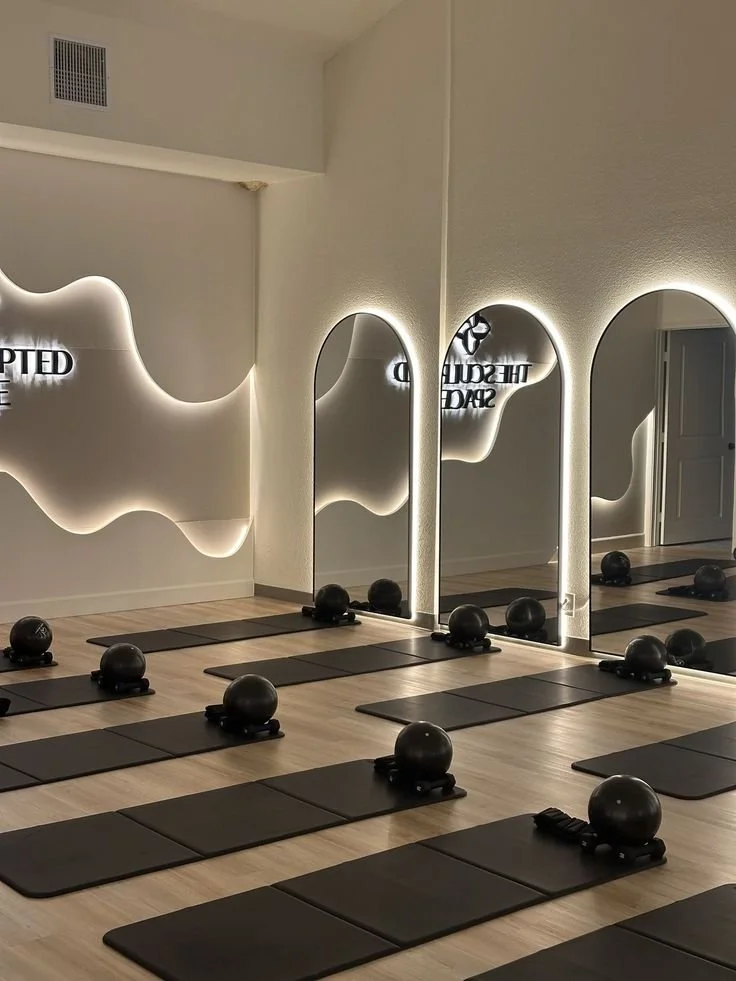 Empty fitness studio with black exercise mats and kettlebells arranged on the floor, mirrors on the wall reflecting the room, and a modern lighted wall art with abstract shapes.
