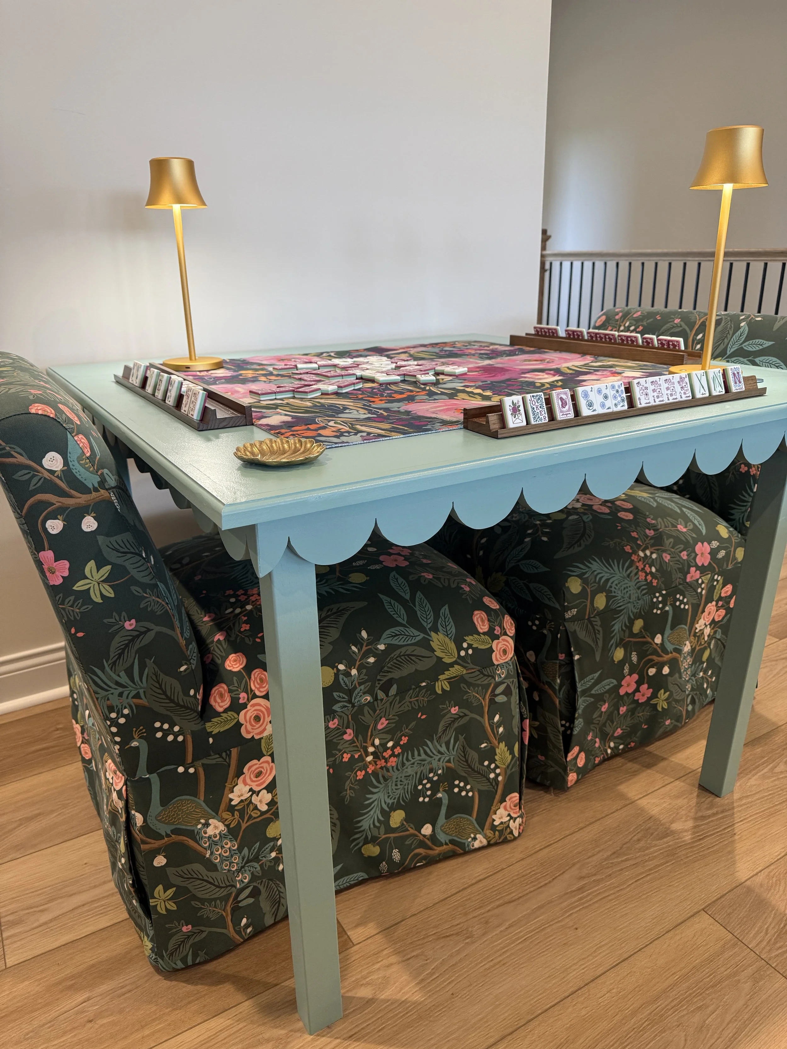 Custom mint green mahjong table with scalloped edges, styled with floral chairs, a floral runner, gold lamps, and mahjong tiles in racks.