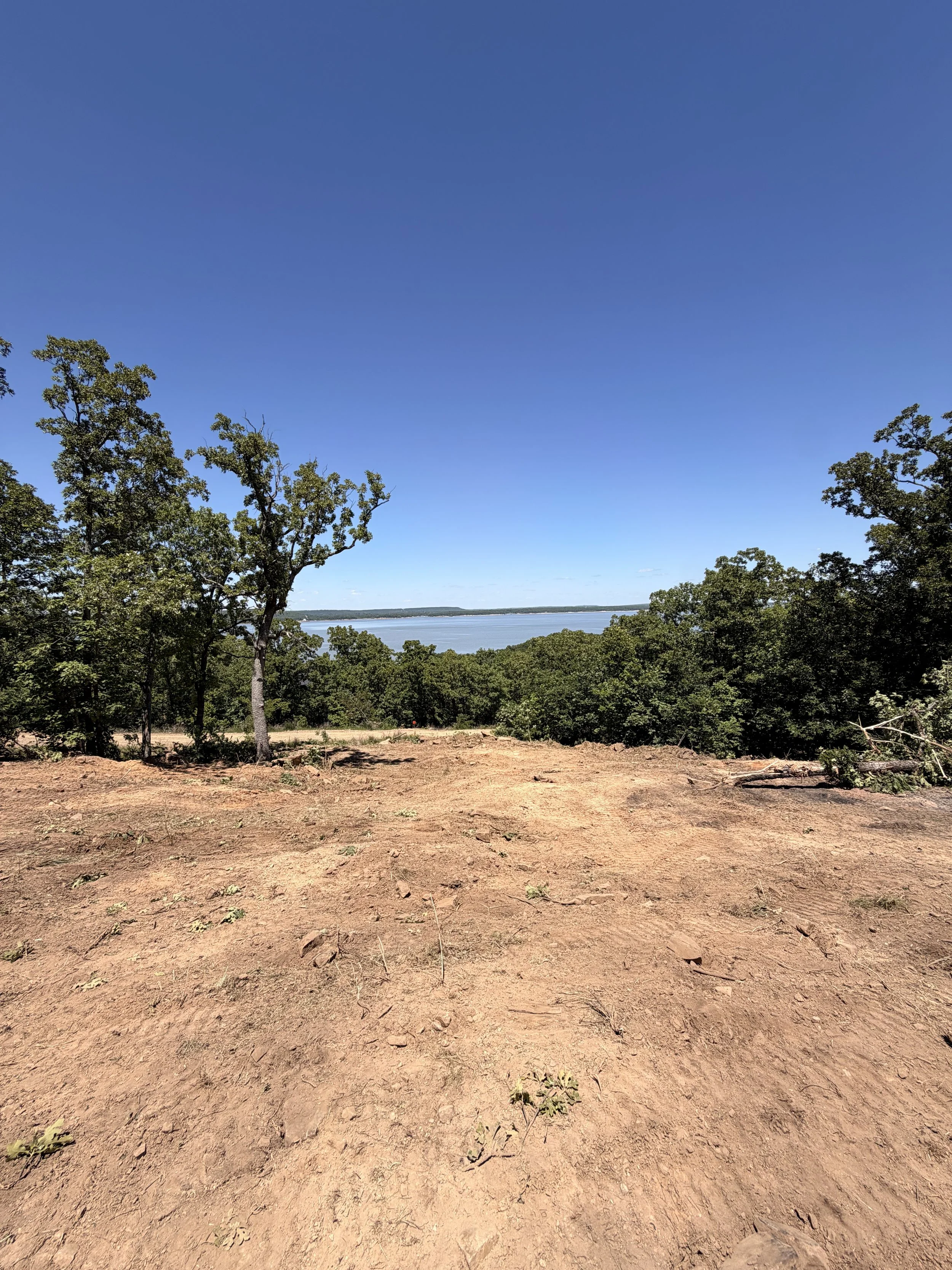 View of a dirt clearing with trees on both sides, overlooking a body of water under a clear blue sky.