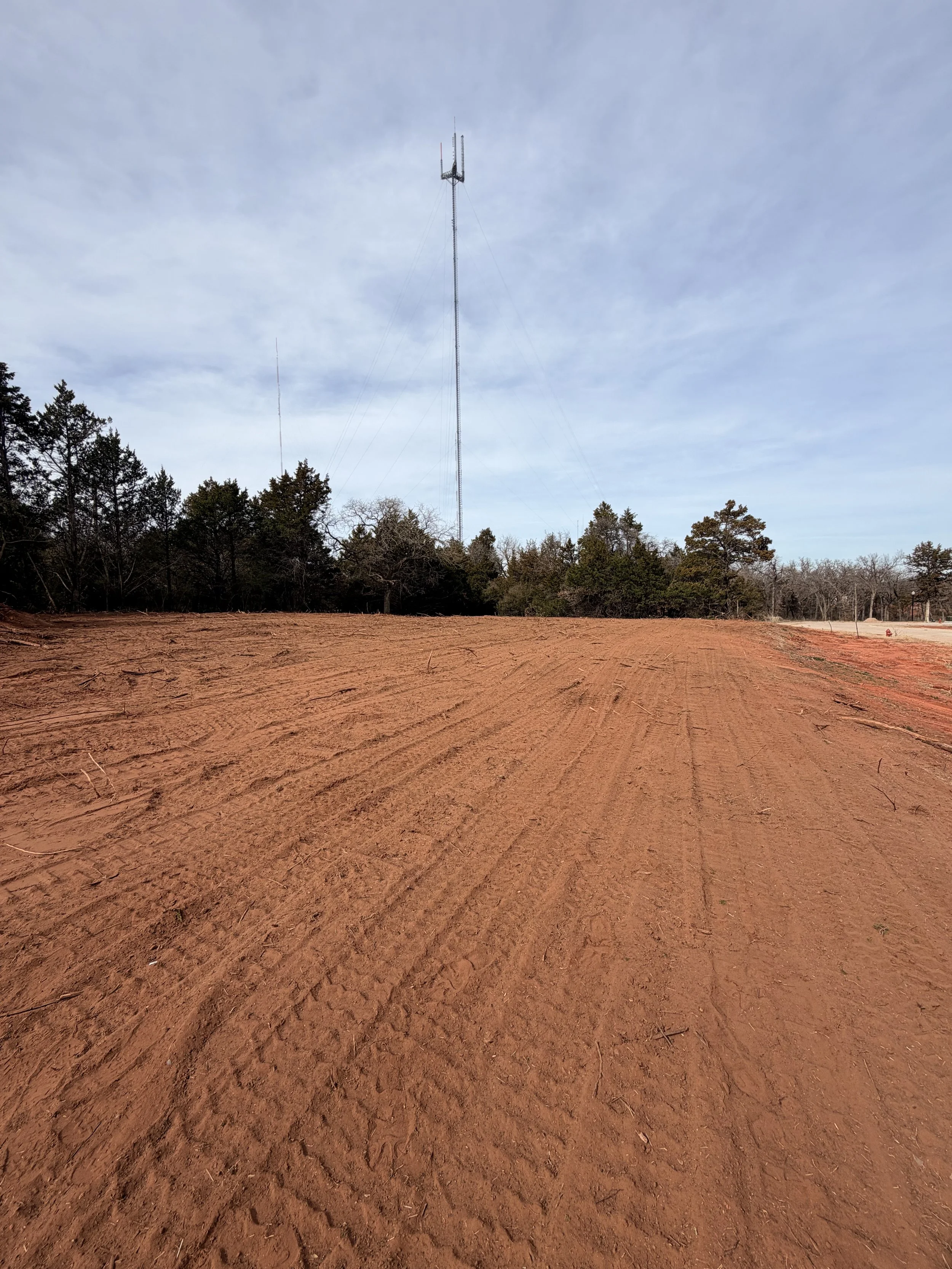 A wide view of a large dirt field with tire tracks, bordered by a row of trees under a blue sky with some clouds, and a tall radio tower in the background.