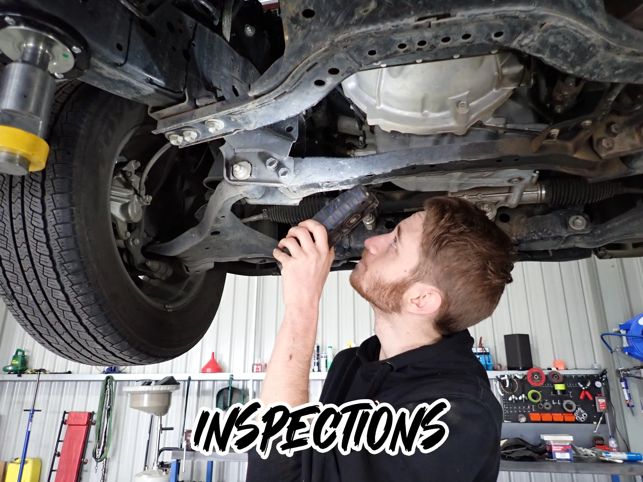 A young man is inspecting the underside of a vehicle raised on a lift in a garage or workshop. The garage is organized with tools and equipment on shelves and pegboards, and the words 'Inspection' are written on the image.