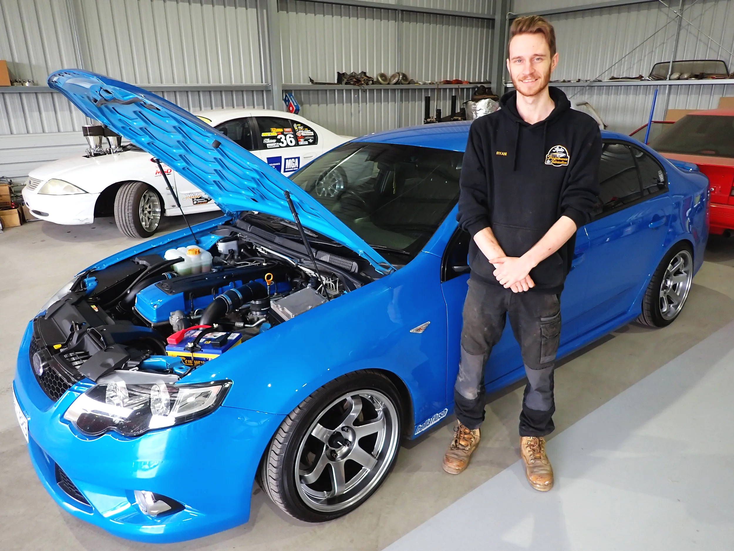 A young man standing in a garage next to a bright blue sports car with its hood open, revealing the engine. There is a white race car with racing decals in the background.