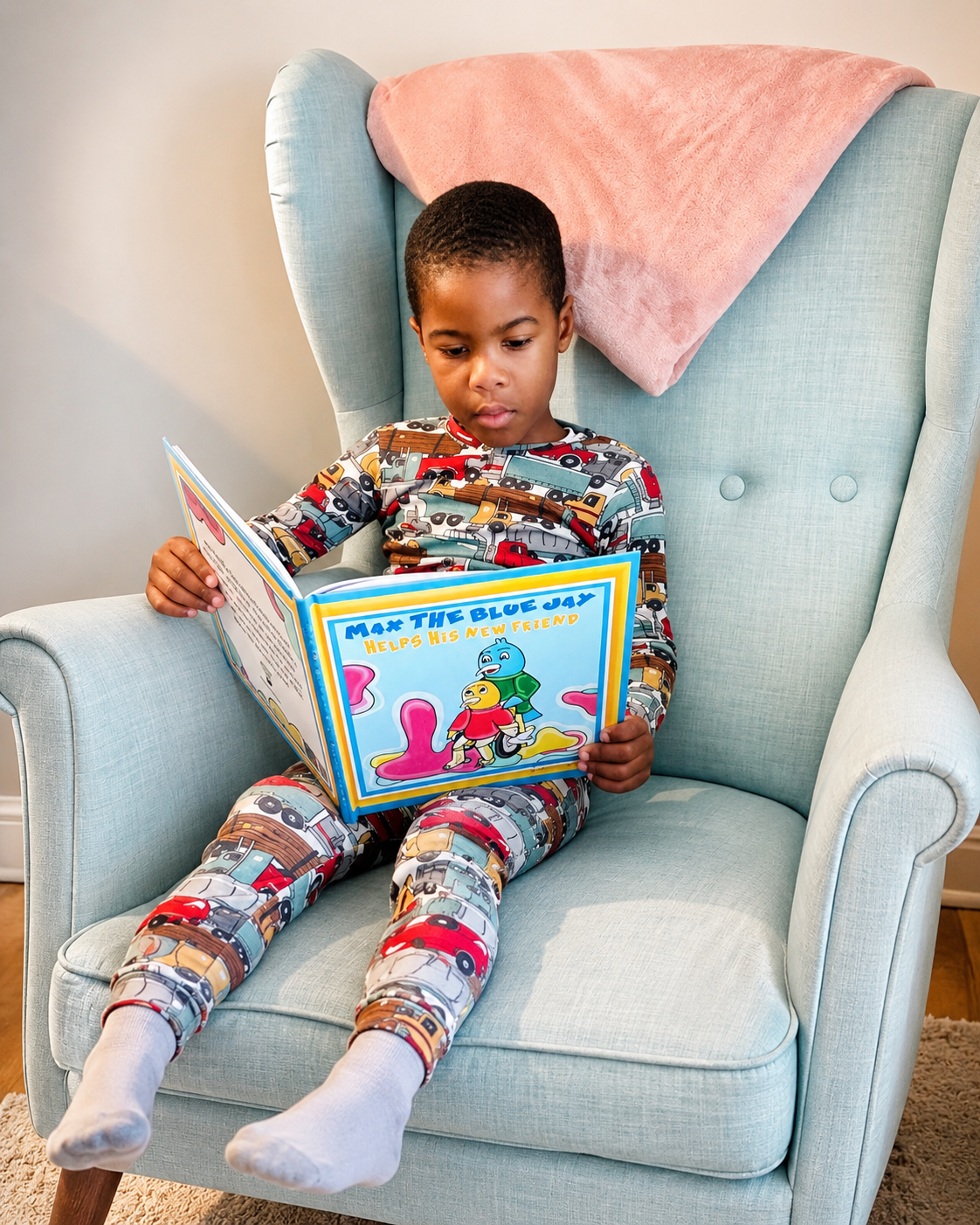 A young boy wearing colorful pajamas with vehicle patterns sits on a light blue armchair, reading a children's book titled "Max the Blue Jay Helps His New Friend." There is a pink blanket draped over the back of the chair.