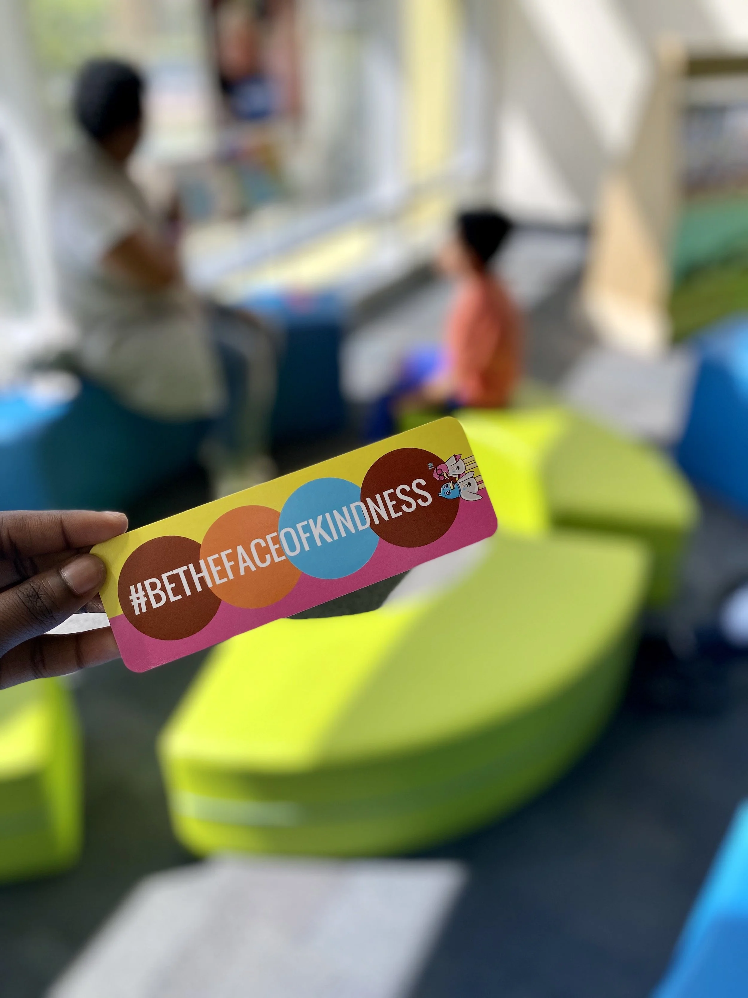 Hand holding a colorful sticker with the hashtag #BETHEFACEOFKINDNESS, showing clouds and a unicorn illustration, with blurred figures in the background and a yellow chair.
