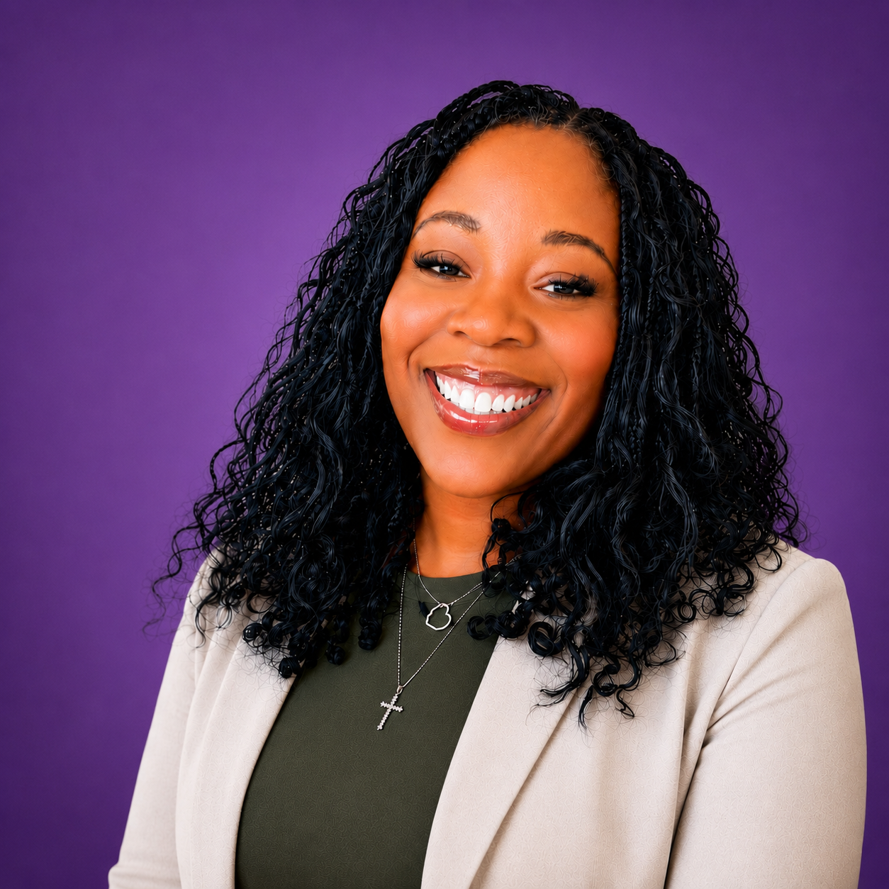 A smiling woman with curly black hair wearing a beige blazer, a dark green top, and silver jewelry, against a purple background.