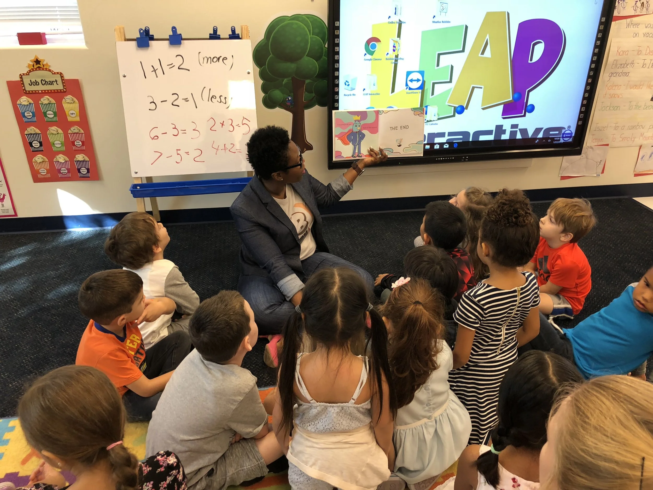 A teacher sits on the floor in front of a group of children, holding a picture book while pointing at an interactive whiteboard displaying the word 'READ!' with colorful letters. Whiteboards with educational content, a tree painting, and students sitting on a colorful rug are visible in the classroom.