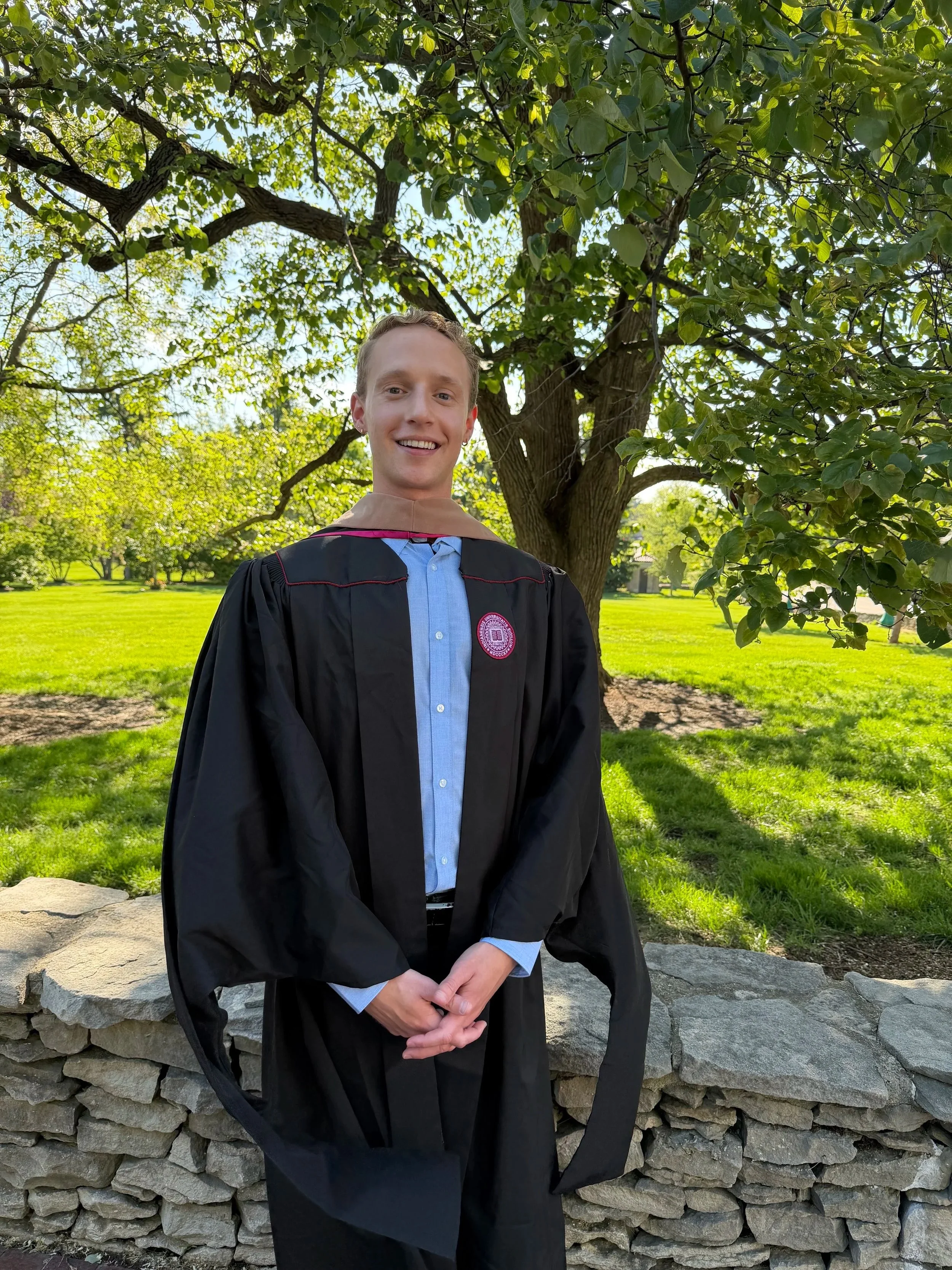 A young man in a black graduation gown and beige hood standing outdoors in front of a stone wall and a large leafy tree, smiling for a photograph.