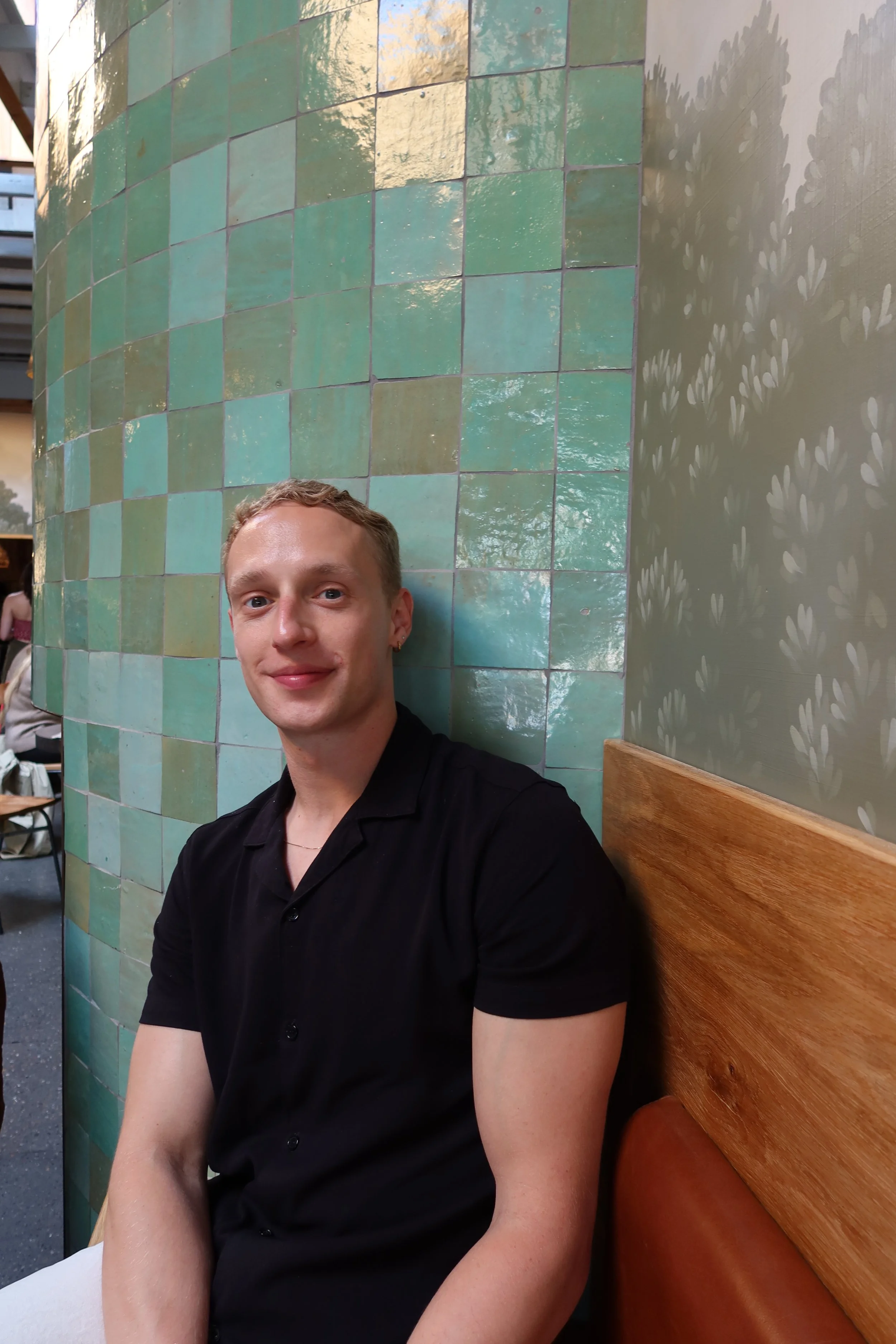 A young man with light brown hair and fair skin, smiling, sitting on a wooden bench against a wall of green tiles, in a room with a patterned wall.