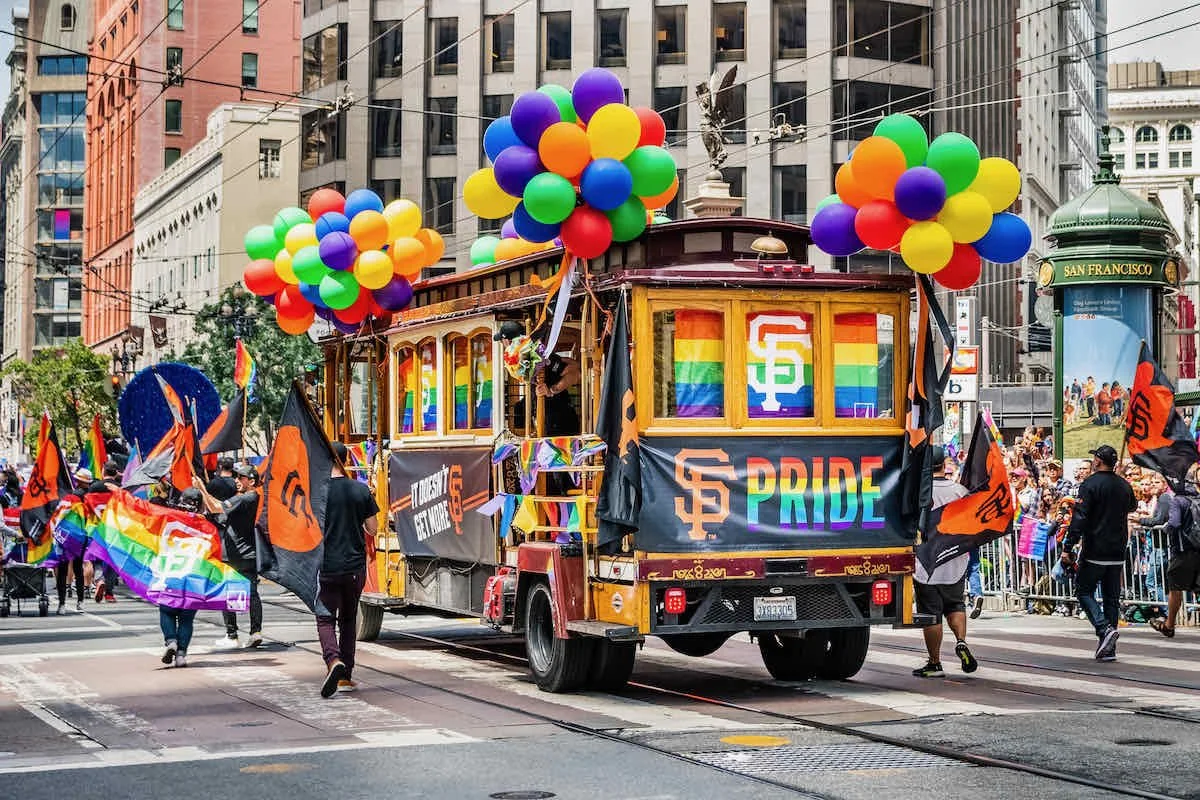A parade float decorated with rainbow pride flags, balloons, and San Francisco Giants branding, celebrating LGBTQ+ Pride in San Francisco with participants holding rainbow flags and walking alongside the float.