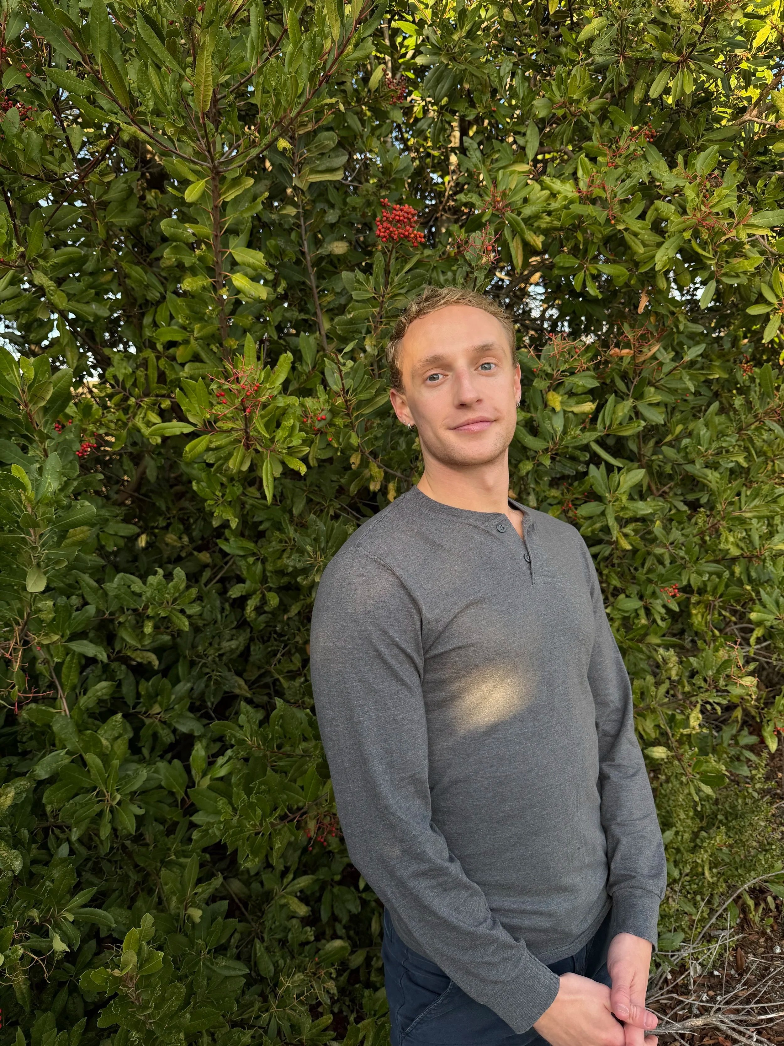 A young man with light skin, blonde hair, and blue eyes is standing in front of a leafy green bush with red berries. He is wearing a gray long-sleeve shirt and dark jeans, and is smiling gently while looking at the camera.