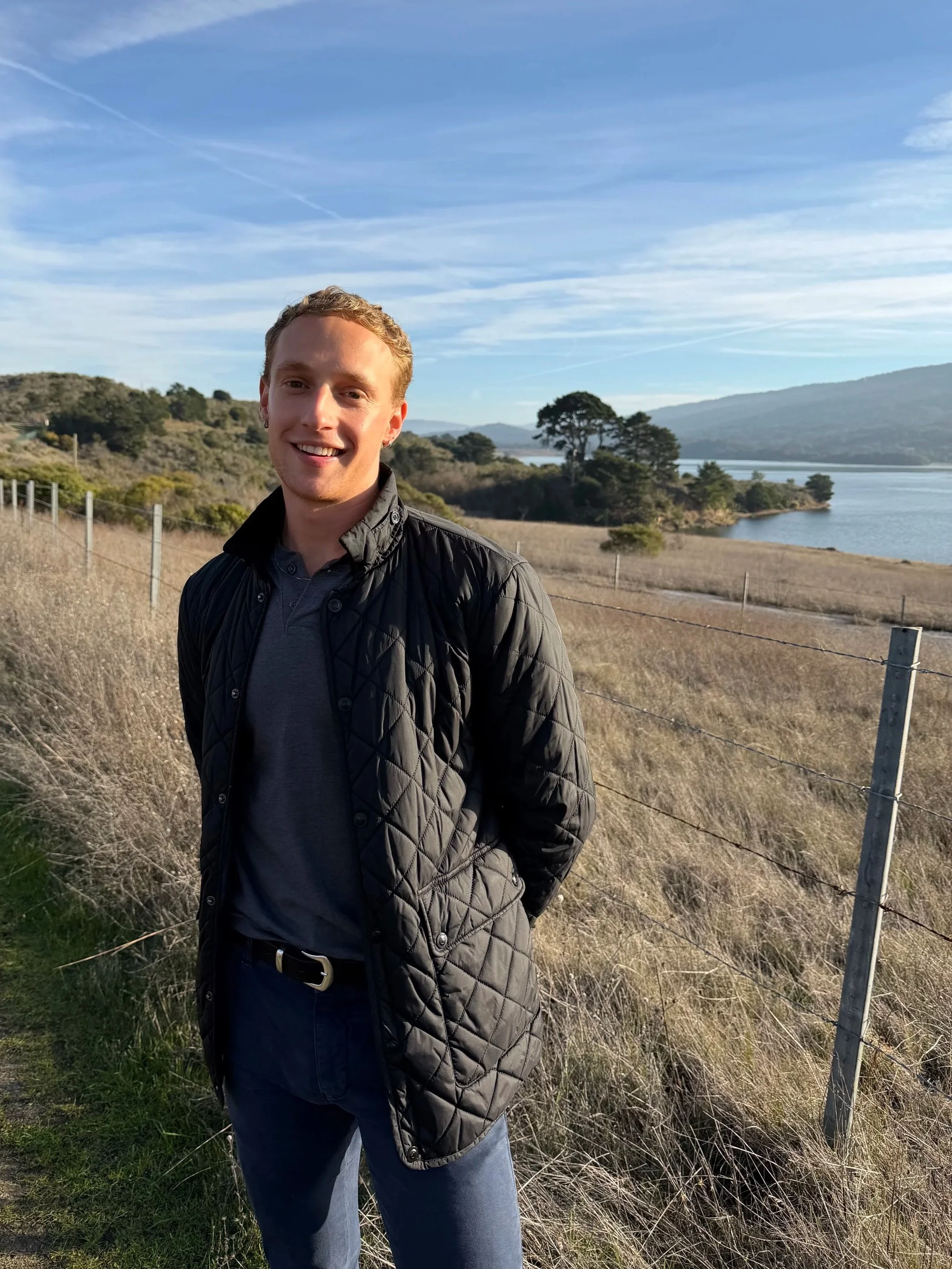A young man standing outdoors near a body of water and green hills, wearing a black quilted jacket and smiling.