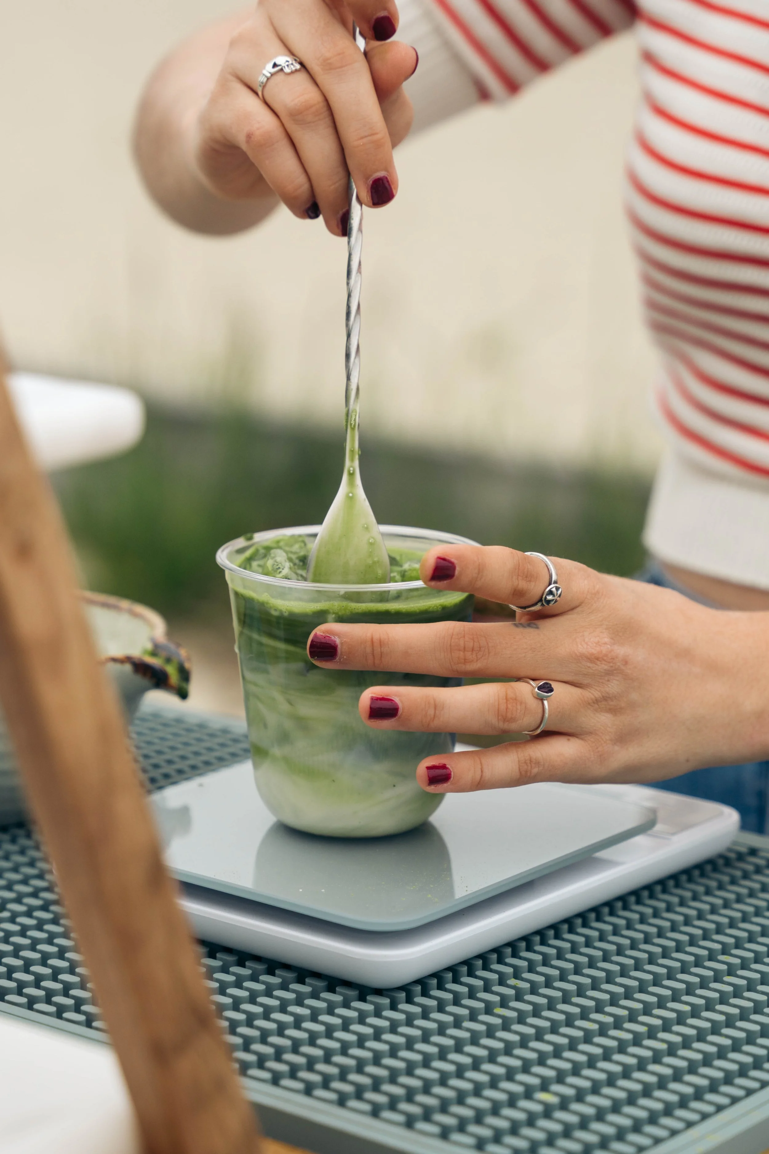 A person with dark red nails stirring a green matcha latte in a clear plastic cup with a metal spoon at a table.