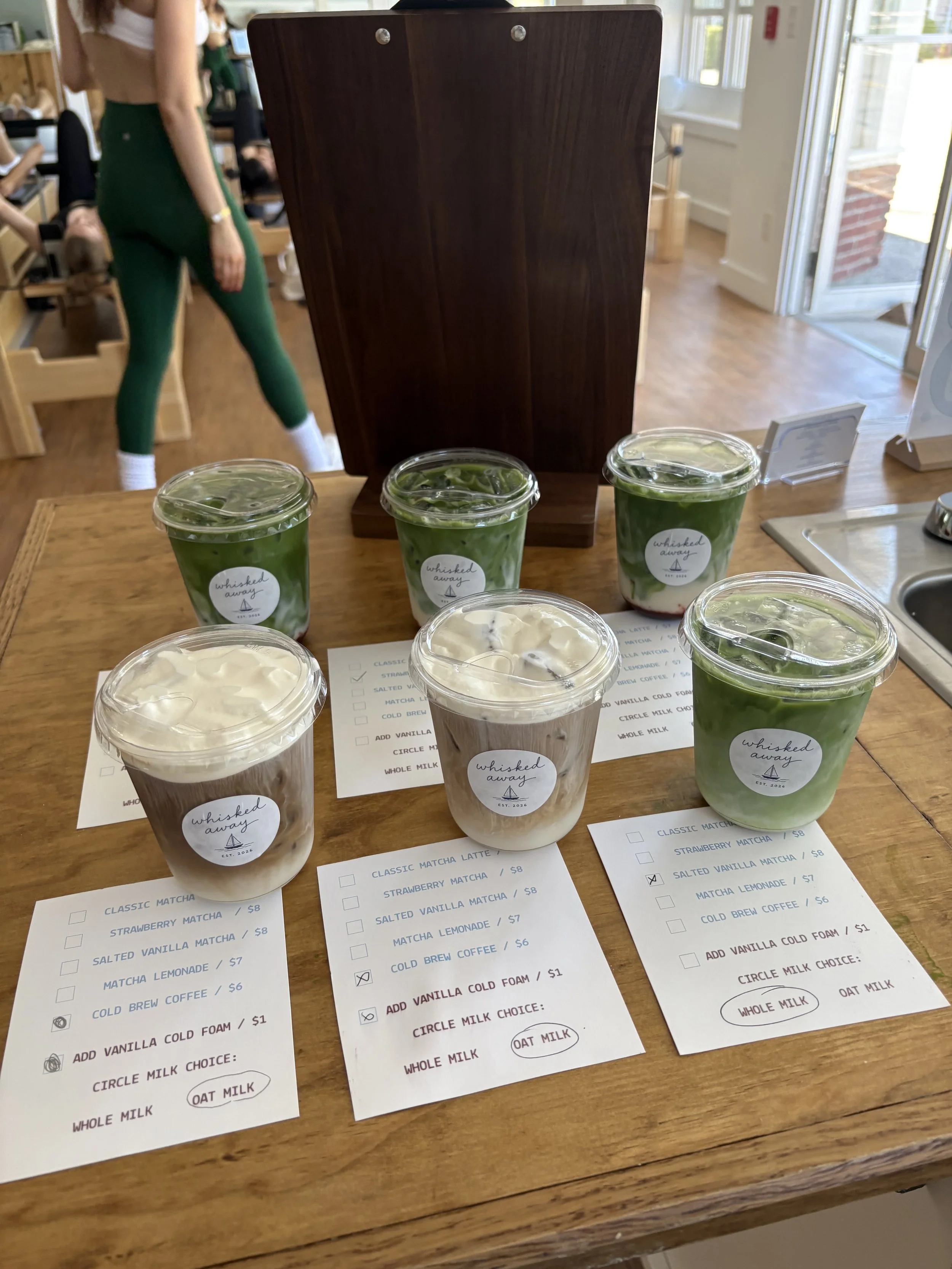 Table in a cafe with several cups of matcha and vanilla drinks with ice, menu sheets, and a dark wooden sign in the background.