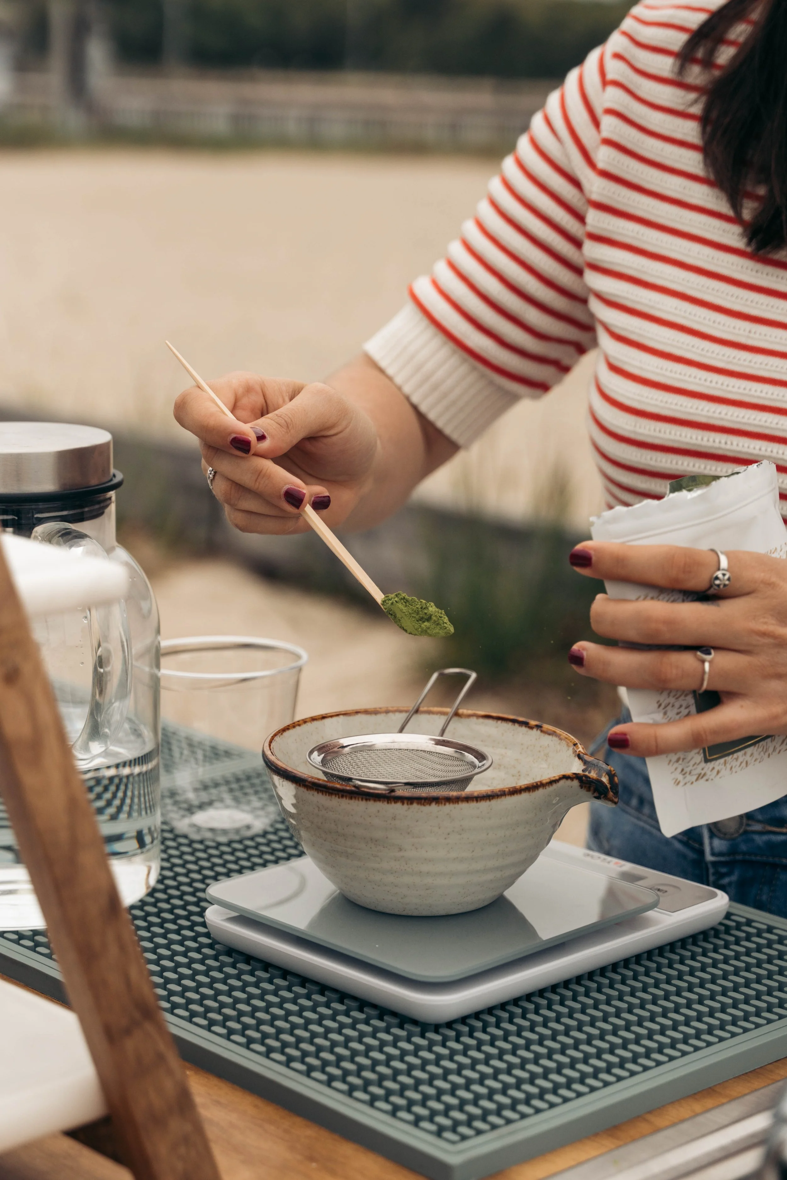 A person is preparing matcha tea, holding a bamboo spoon with matcha powder over a ceramic bowl on a scale. Various items including a glass cream pitcher, a spoon, and a bag of matcha are on the table.