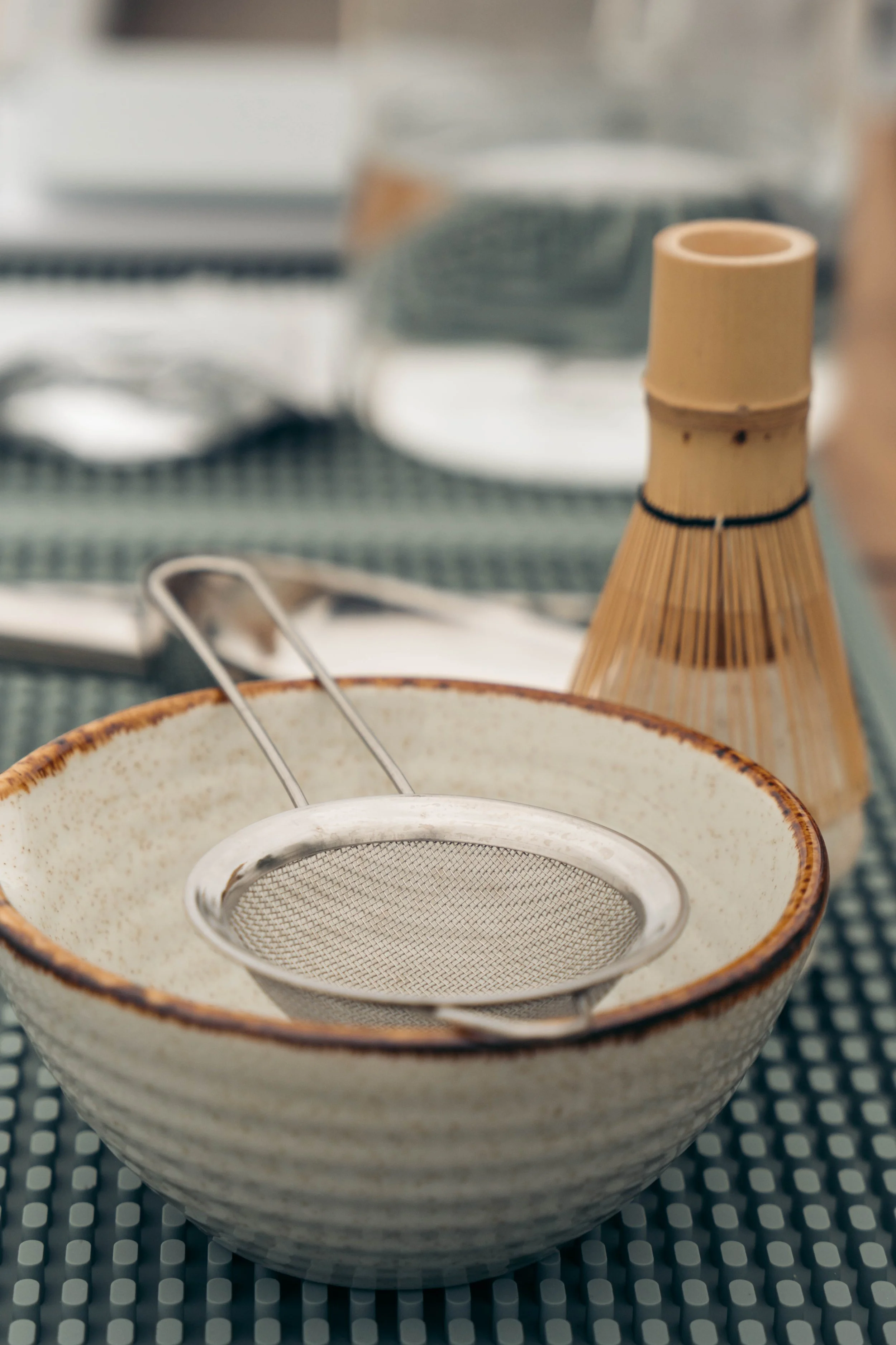 A ceramic bowl with a brown rim, containing a metal mesh strainer, in front of a blurry background with kitchen items, including a small bamboo bottle and a dish drying rack.
