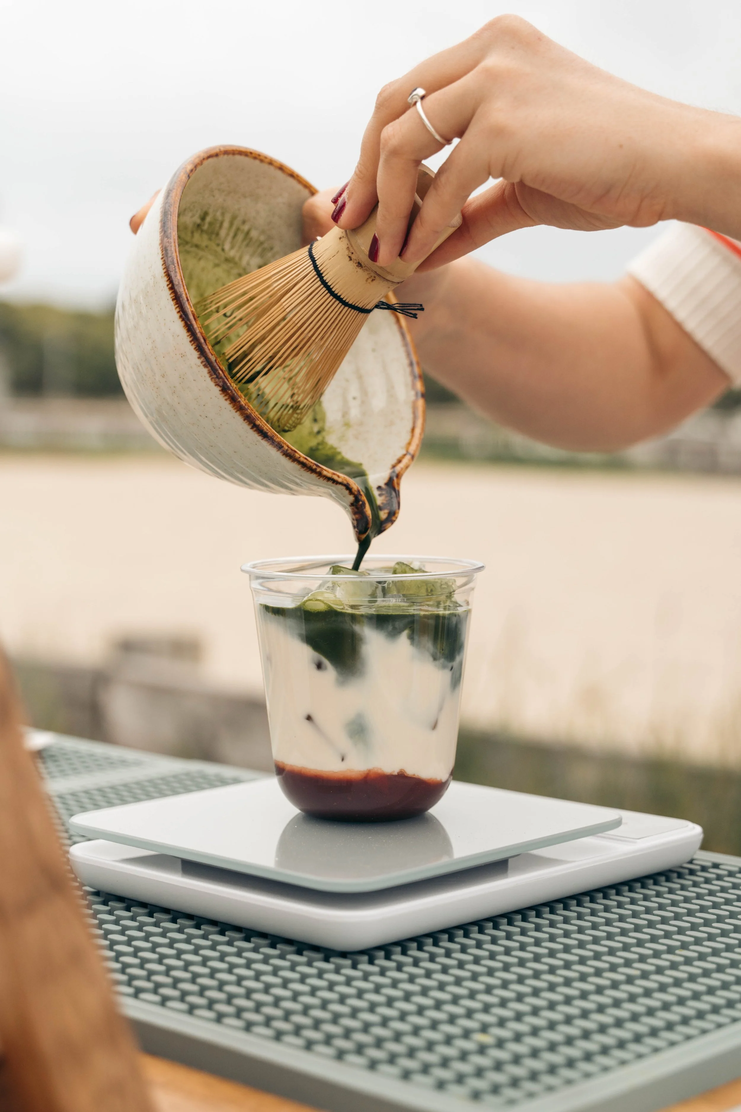 A person using a bamboo spoon to serve green matcha or tea from a ceramic teapot into a clear glass filled with ice and layered dessert. The scene is outdoors, on a table with a scale underneath the glass.