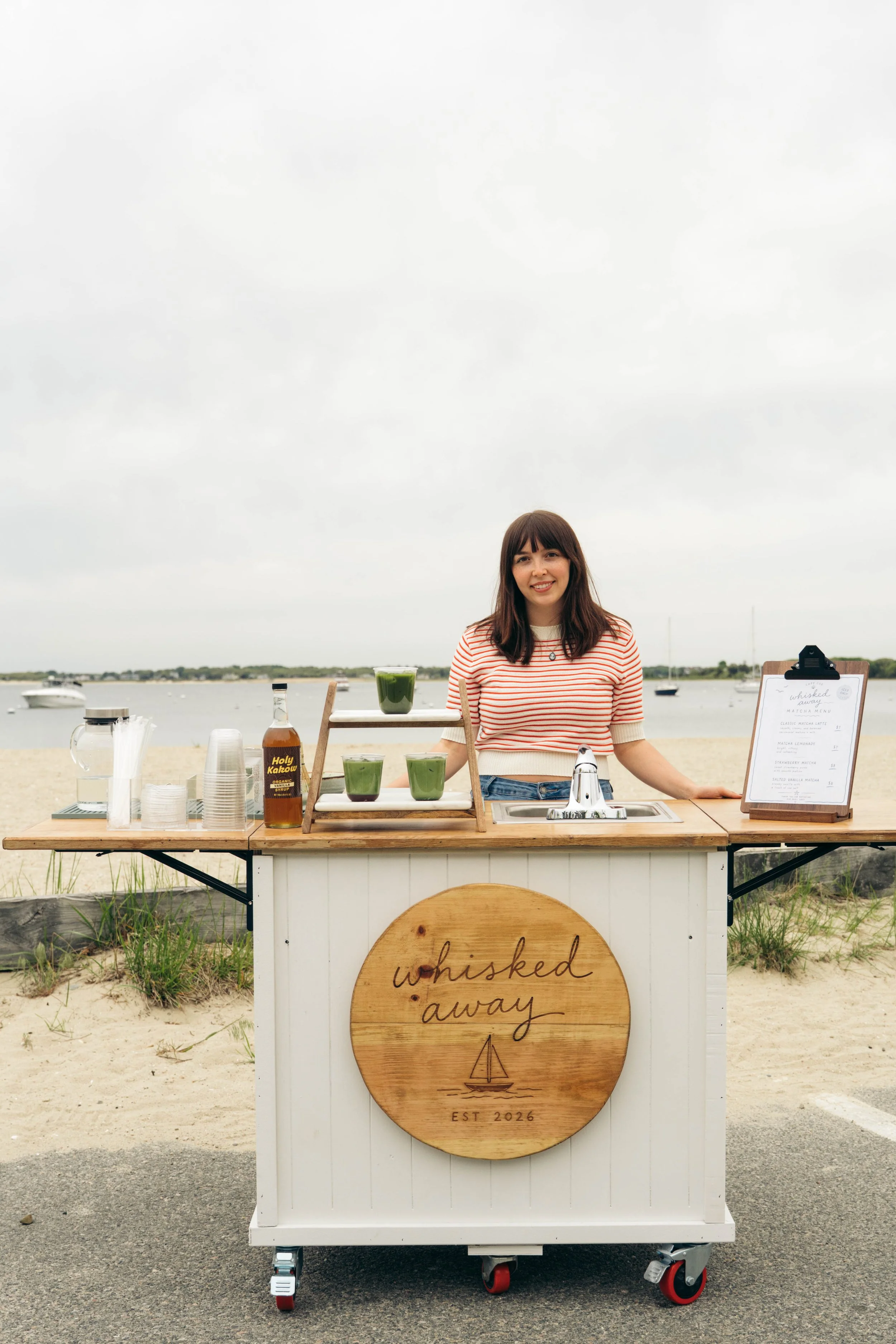 A smiling woman with dark hair standing behind a white outdoor drink cart with a round wooden sign that reads "whisked away" and features a sailboat illustration, set against a cloudy sky over a body of water with boats in the background.