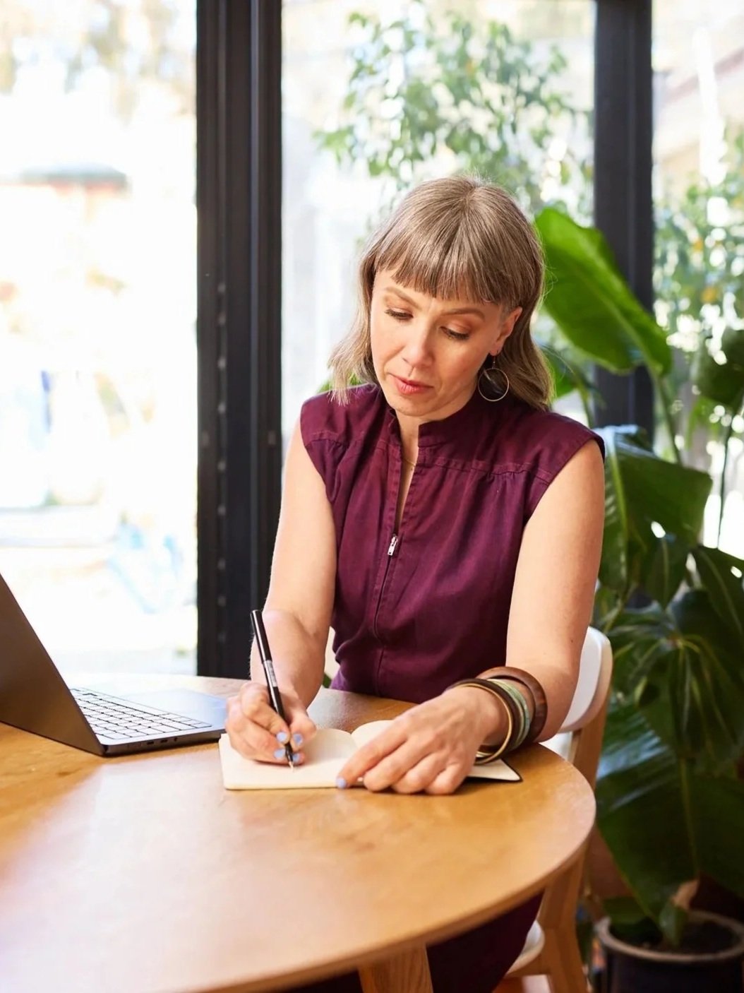 A woman sitting at a wooden table, writing in a notebook with a pen. There is a laptop beside her, and large green plants behind her.