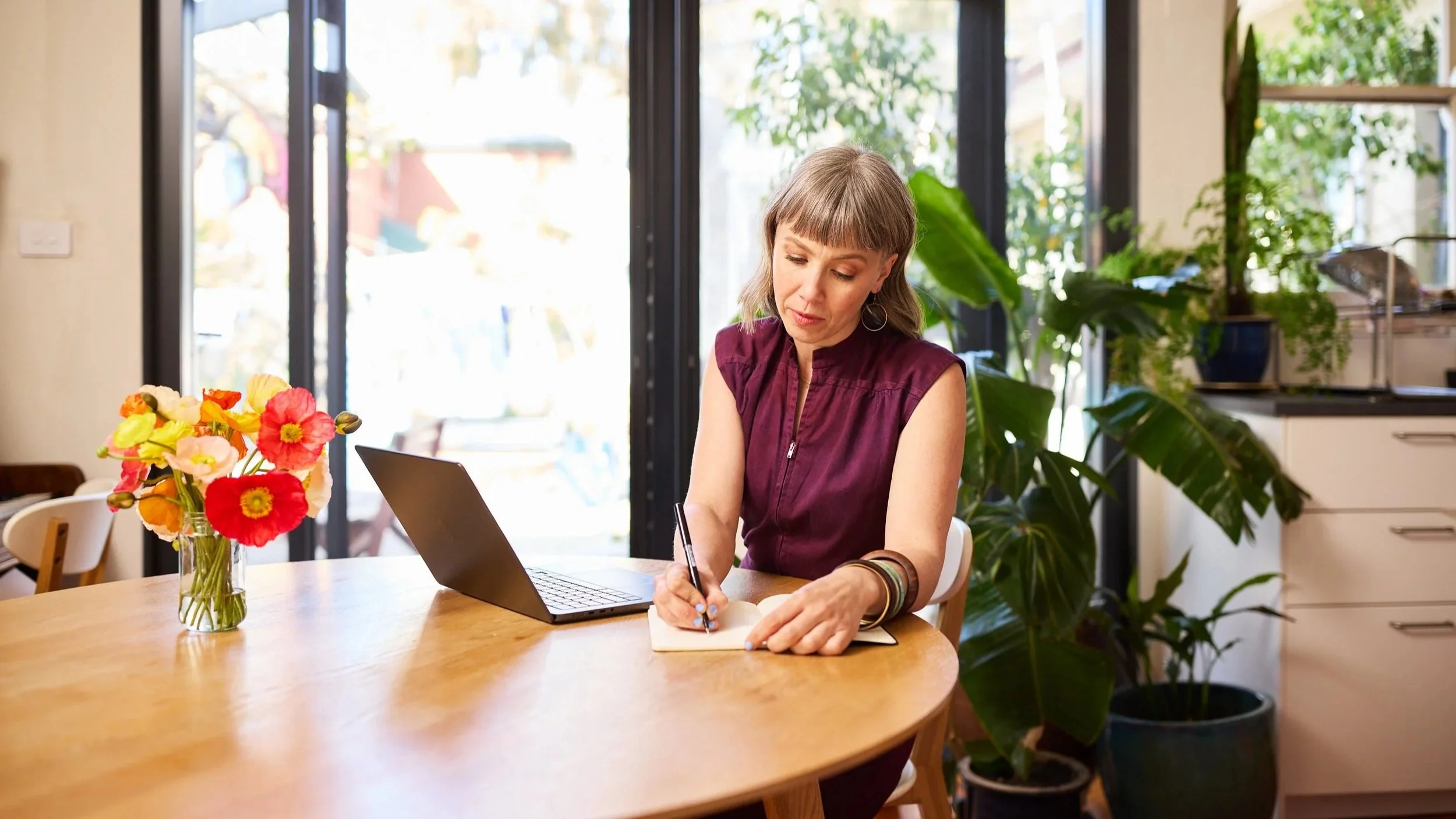Koral - founder of Moon Mode and Menstruality Mentor for leaders - sits at a wooden dining table, writing in a notebook with a pen, with a laptop open in front of her.