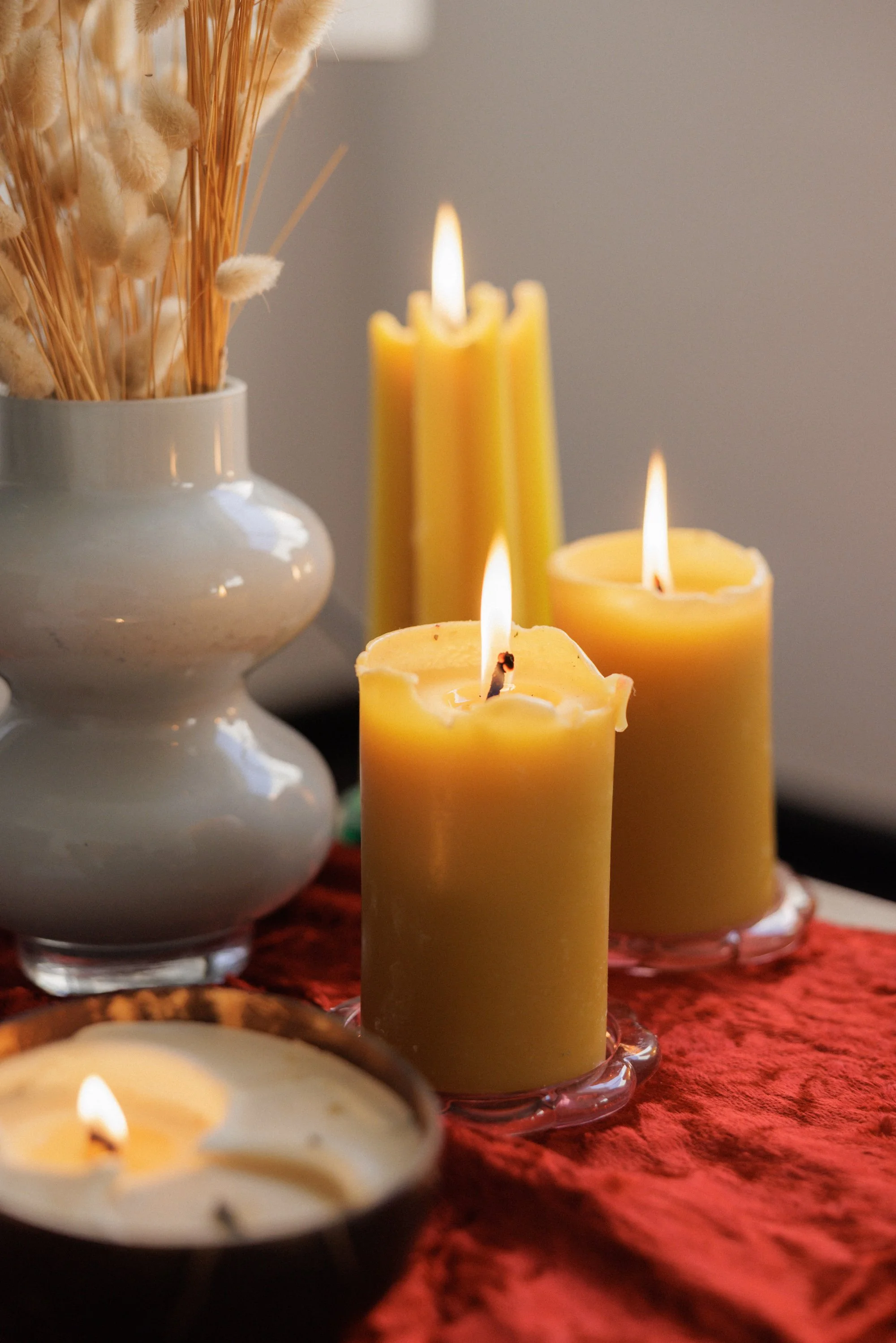 Multiple yellow candles burning on a red textured surface, a white vase with dried pampas grass in the background.