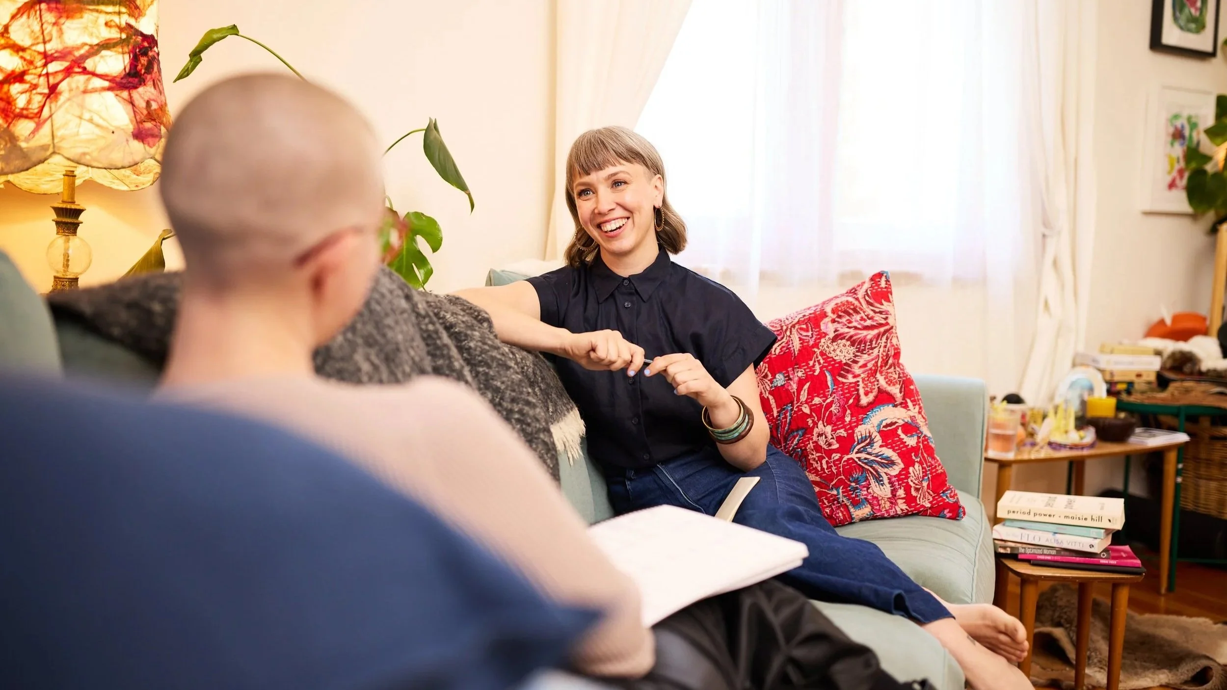 Koral from Moon Mode- smiling and wearing earrings is sitting on a couch, engaged in a conversation with a woman in a 1:1 menstruality mentoring session.