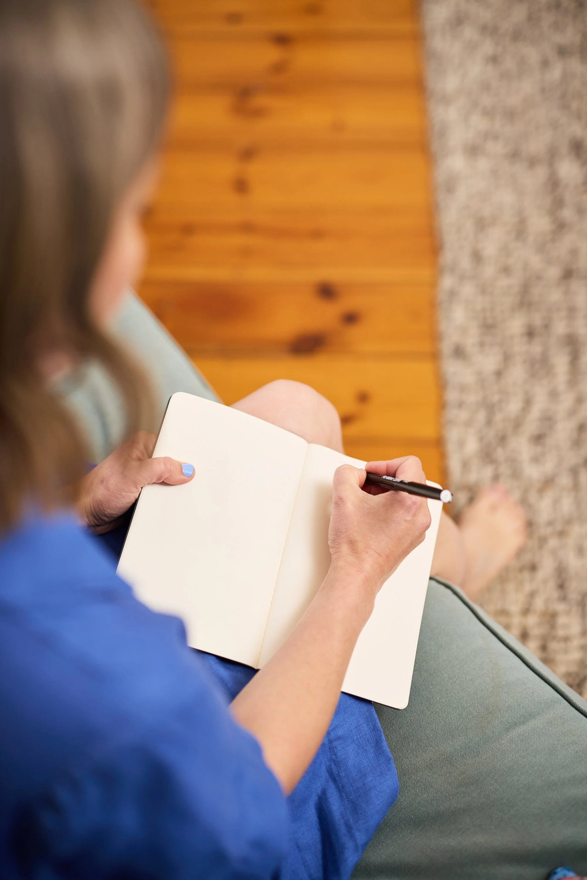 Person sitting on a couch, holding an open notebook and writing with a black pen, wearing a blue shirt, with a wooden wall and carpeted floor in the background.