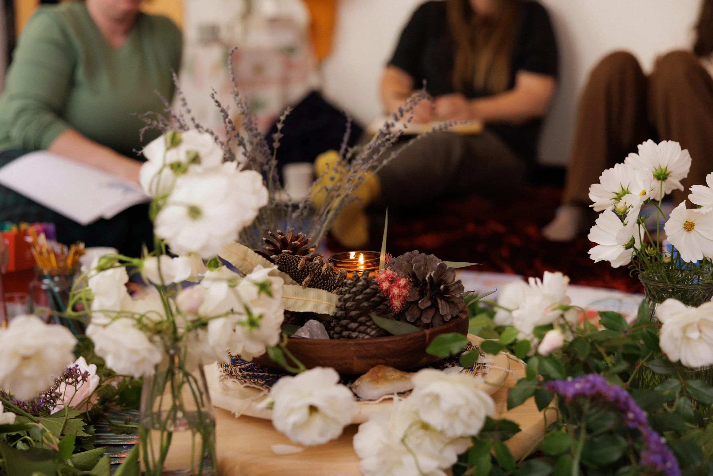 Indoor scene with various white and purple flowers, a lit candle, pinecones, and other decorative items on a table, with two people in the background, one wearing a green shirt and the other in black, engaged in conversation or activity.