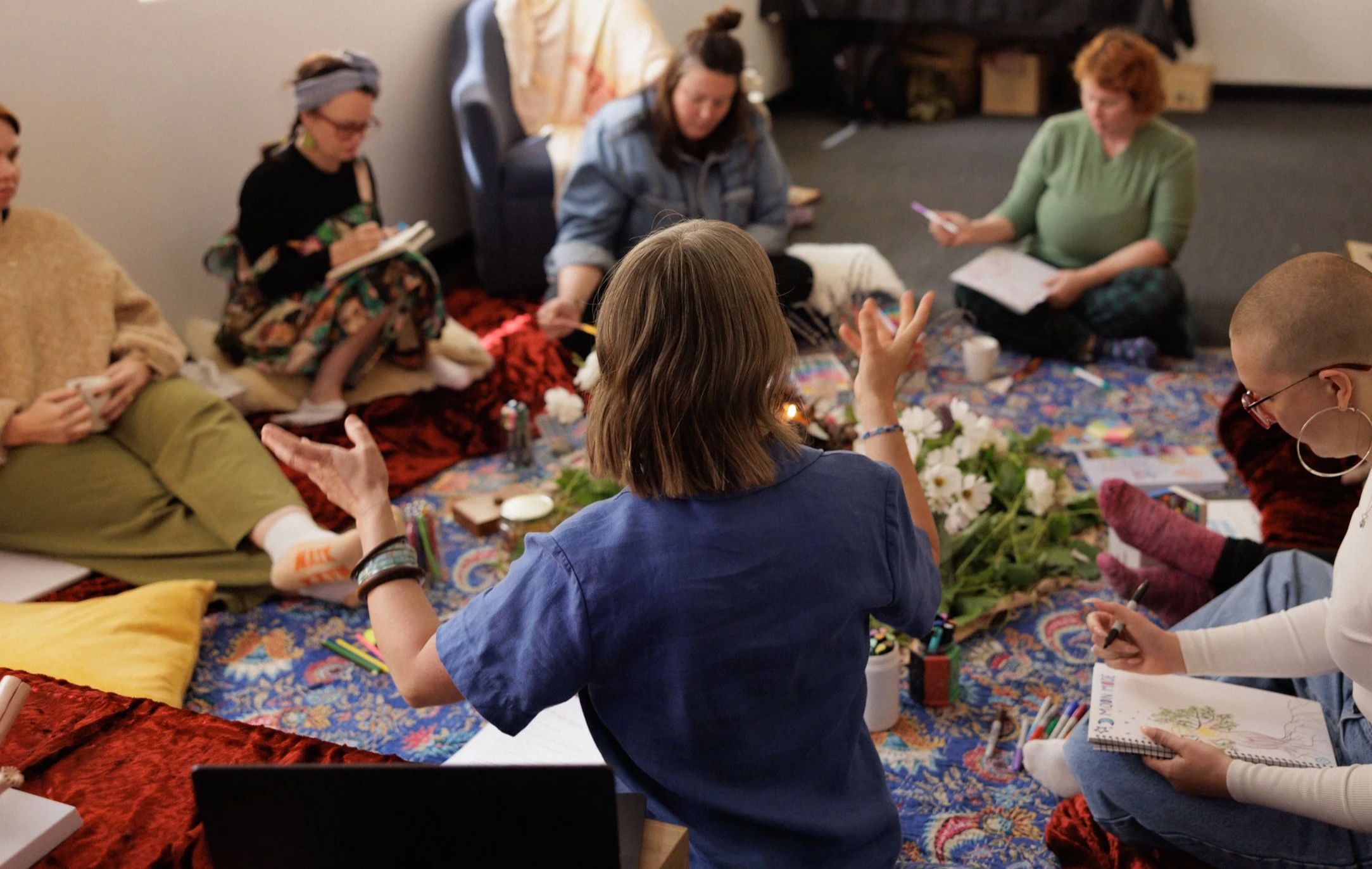 Group of people sitting on a colorful rug, engaged in a Moon mode Foundations of Menstruality workshop, with notebooks, pens, and flowers around them in Adelaide Australia