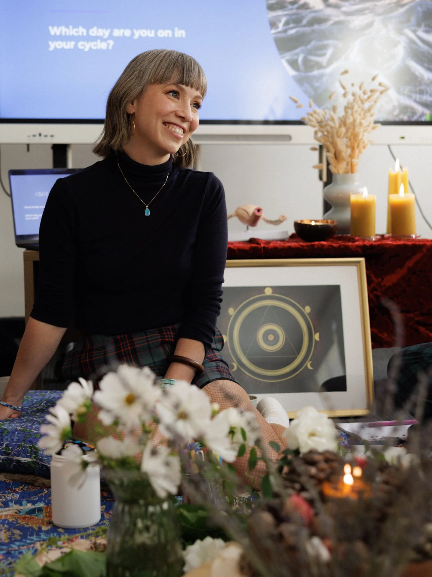 Koral - Moon Mode founder and menstruality mentor - smiling, wearing a black turtleneck and plaid skirt, sitting at a table with flowers and candles, facilitating a workshop