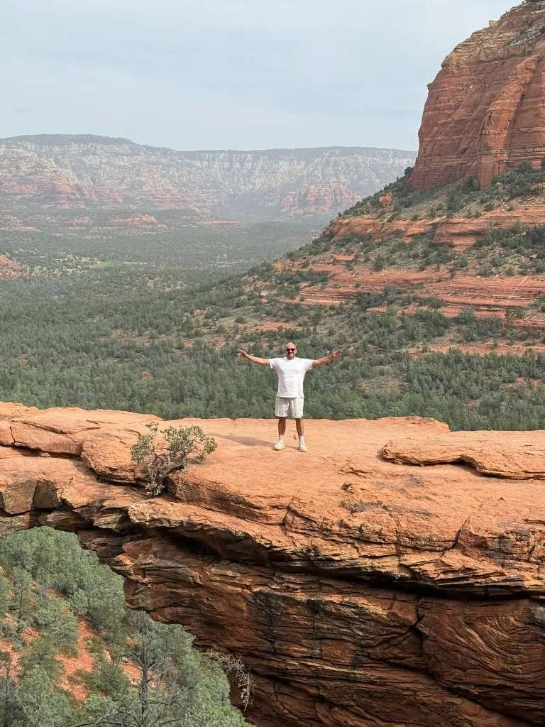A man standing on a narrow rock ledge with arms outstretched, surrounded by red rock formations and a green valley in a desert landscape.