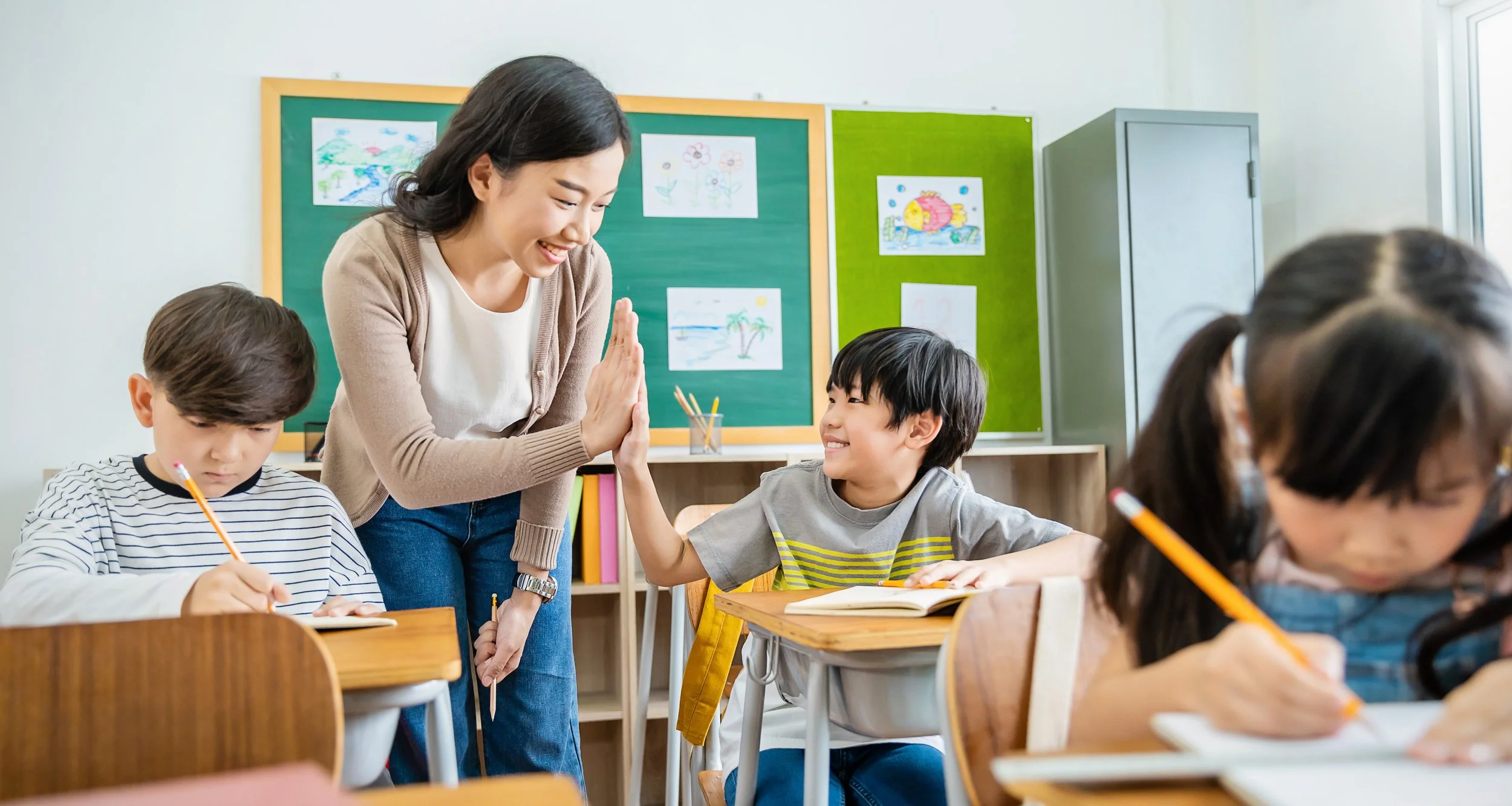 A teacher and students in a classroom, with the teacher smiling and giving a high-five to a student while others work on assignments at their desks.
