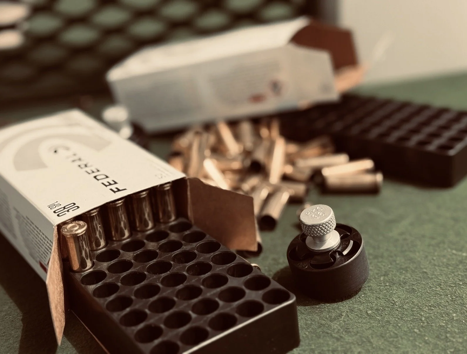 A box of Federal 308 Winchester ammunition with several bullets spilled out, a gun suppressor, and a tray of empty bullet casings on a green surface.