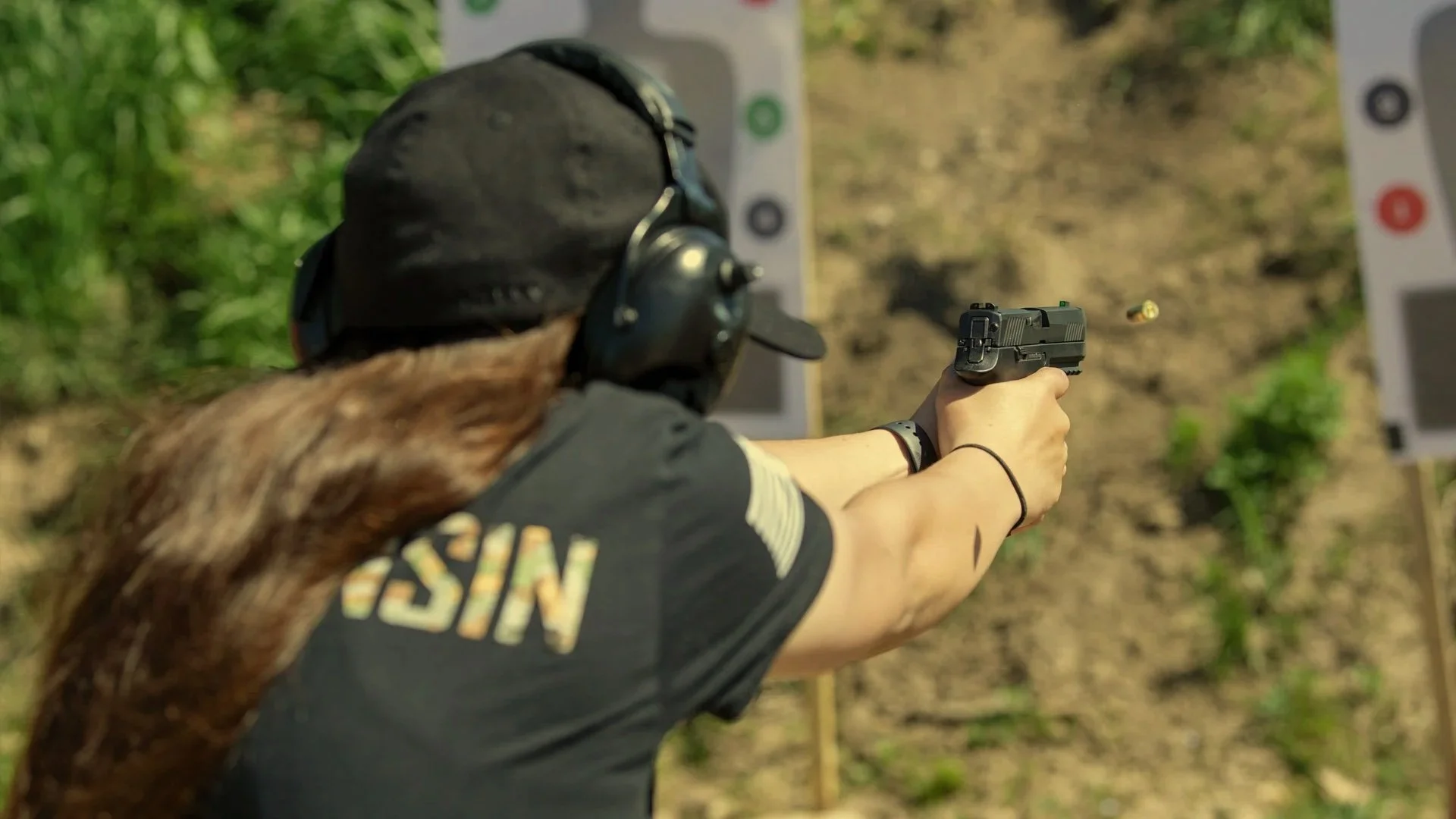 Person shooting a handgun at a target outdoors, wearing ear protection and a tactical vest.