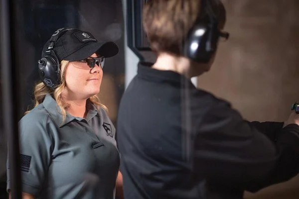 A woman and a man at a shooting range, both wearing ear protection and the woman wearing sunglasses, with the woman observing as the man aims a firearm.