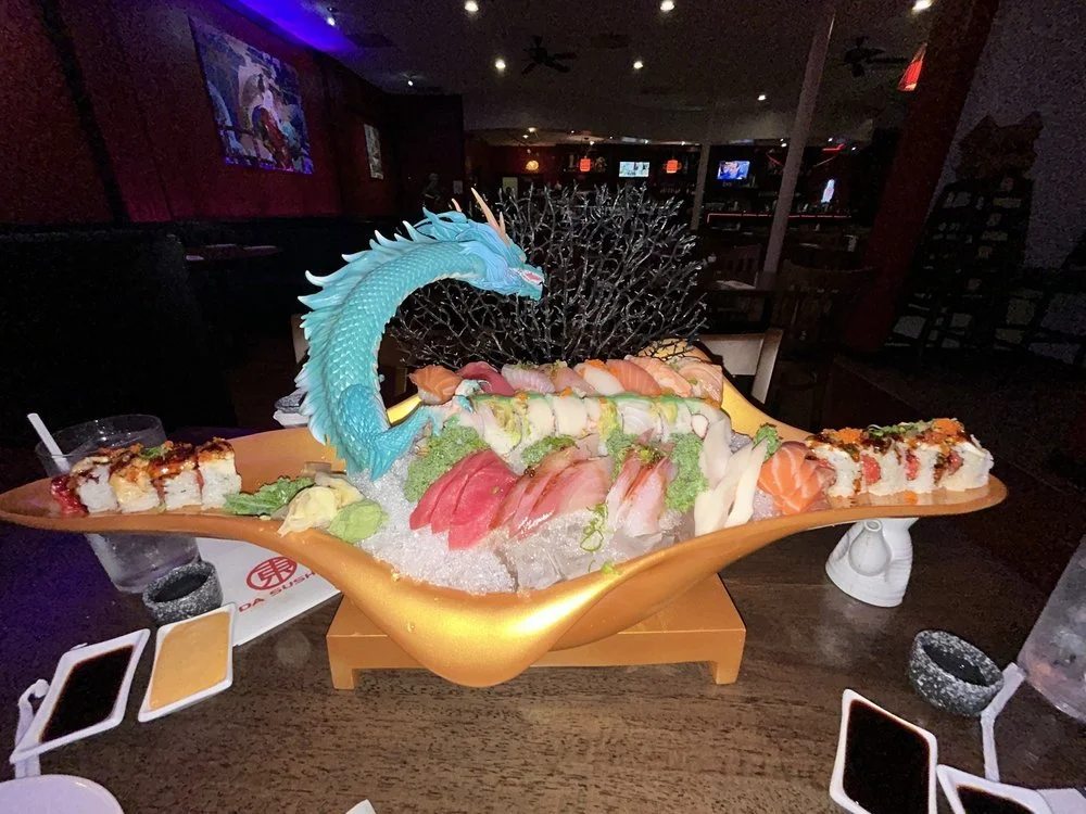 Large sushi platter on a decorative boat-shaped platter with a blue dragon figure, assorted sushi and sashimi, wasabi, and garnishes, set on a dark restaurant table.