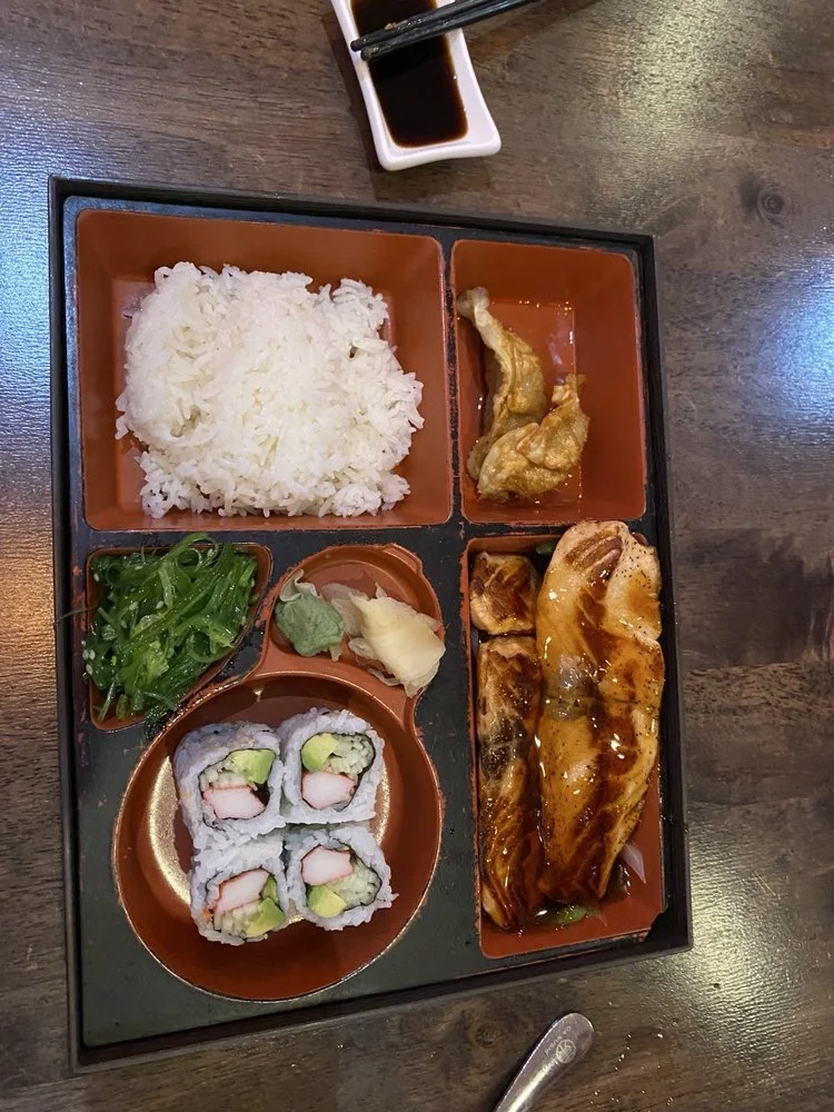 A Japanese bento box with white rice, tempura, seaweed salad, sushi rolls with crab and avocado, sliced eel with sauce, and wasabi and ginger, along with a small container of soy sauce and chopsticks.