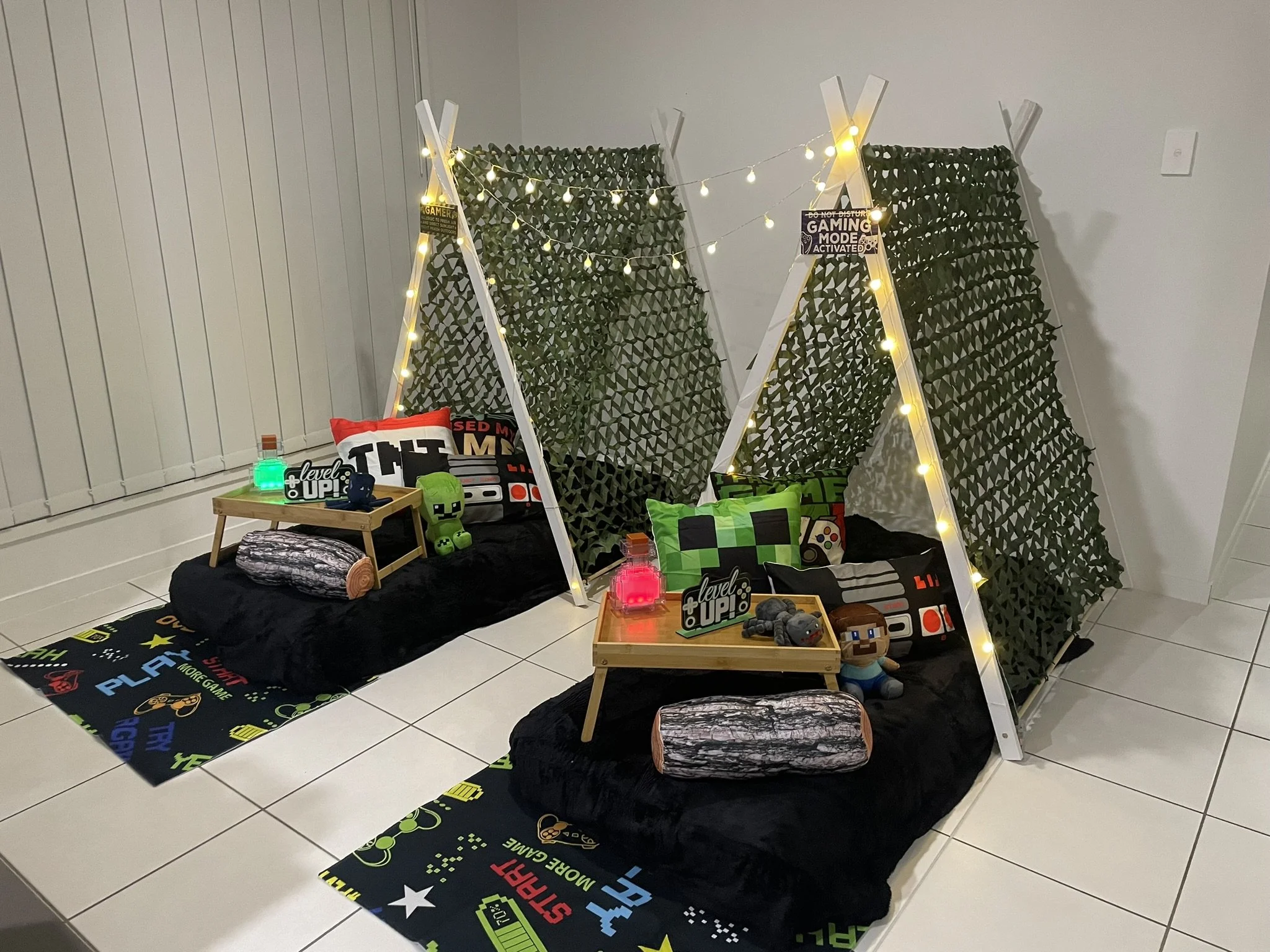 A gaming-themed bedroom setup with fabric panels and string lights forming a tent, decorated with gaming pillows, plush toys, and a gaming-themed rug on white tiled flooring.