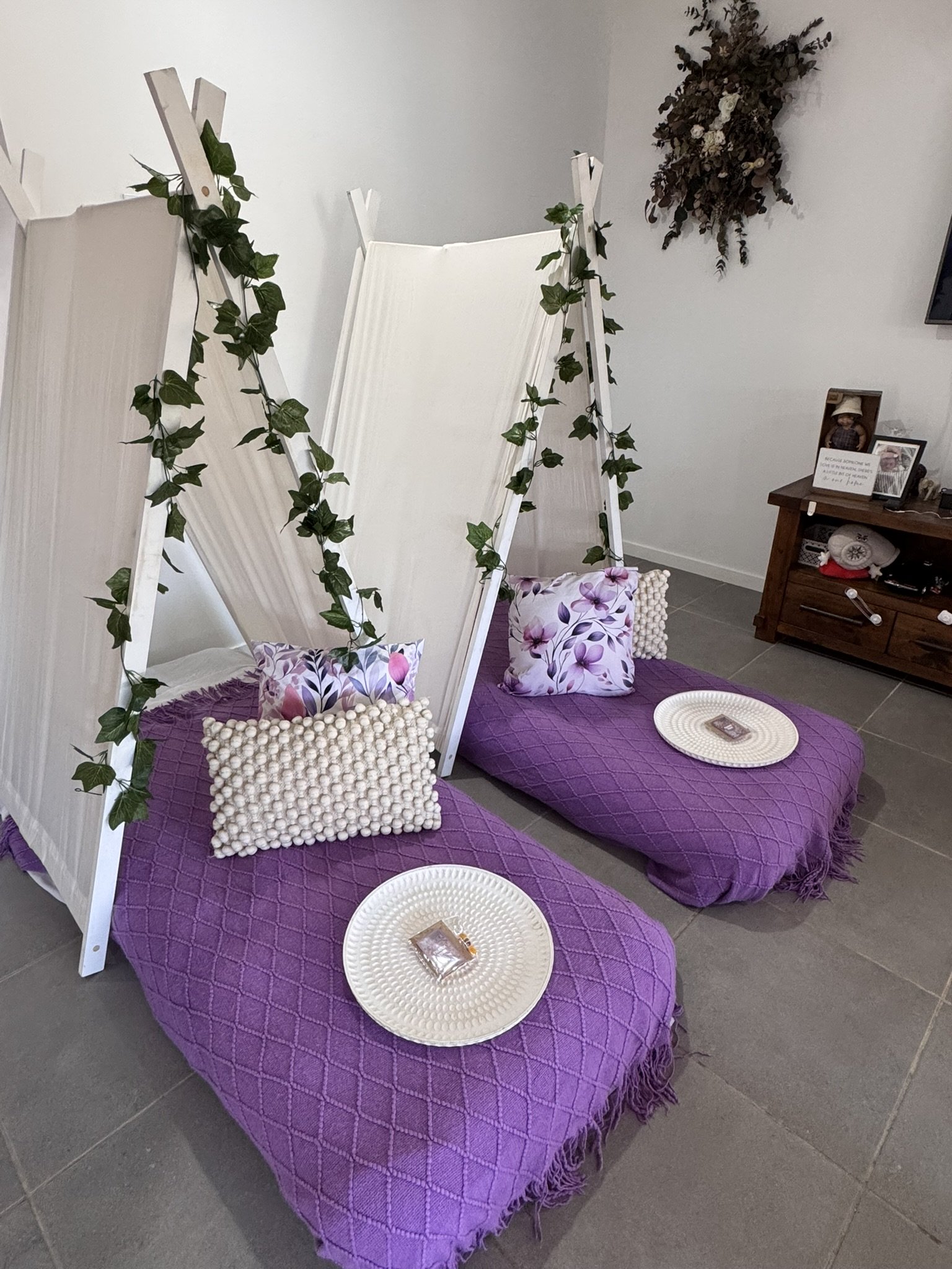 Two A Frame Teepees with purple quilted blankets, pillows with floral patterns, small round white tables, and decorative ivy accents, set up indoors.
