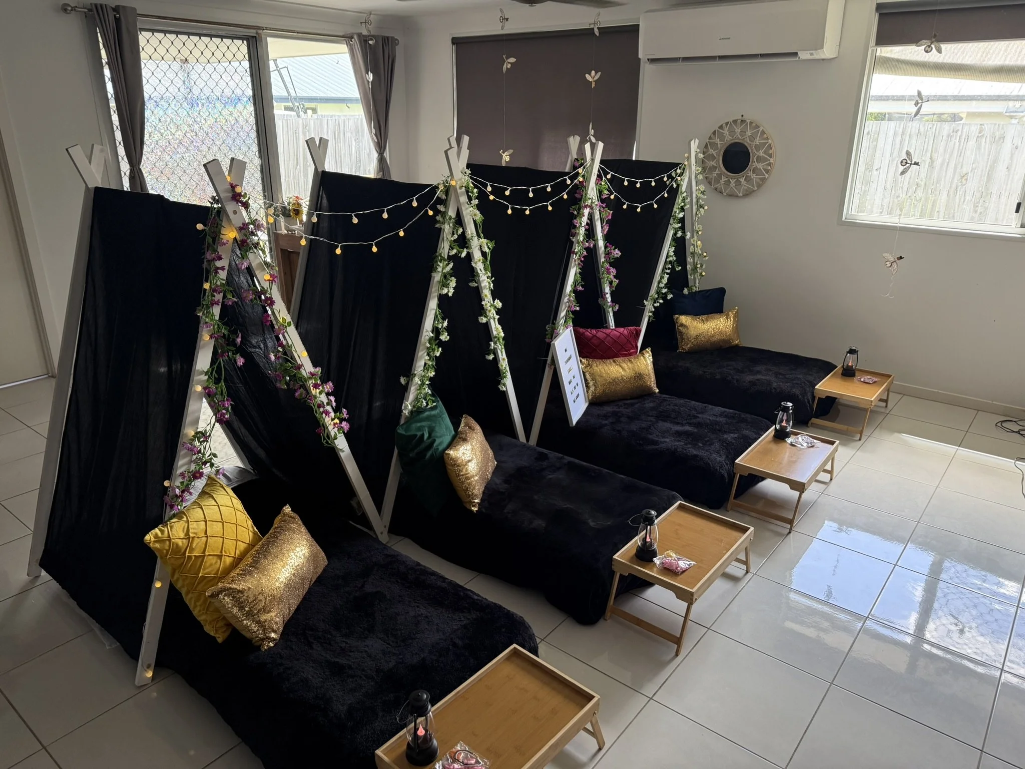 Indoor space decorated with black fabric booths with flower garlands and string lights, gold pillows on black couches, small wooden tables with lanterns, and butterfly decorations hanging from the ceiling window curtains.