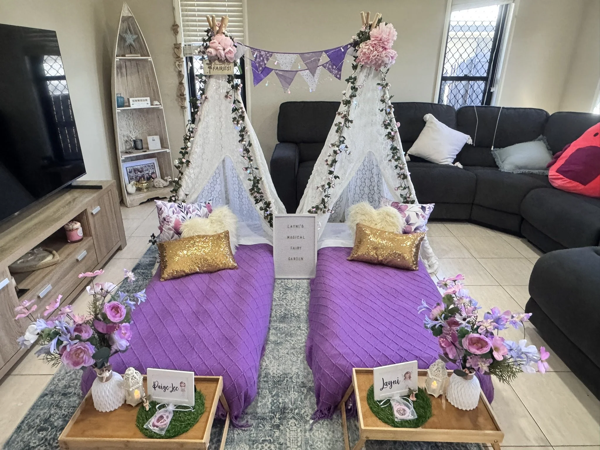 Decorative display for fairy-themed event with two small tent-like structures, pink flowers, fairy garden signs, and fairy figurines surrounded by purple and gold accents.