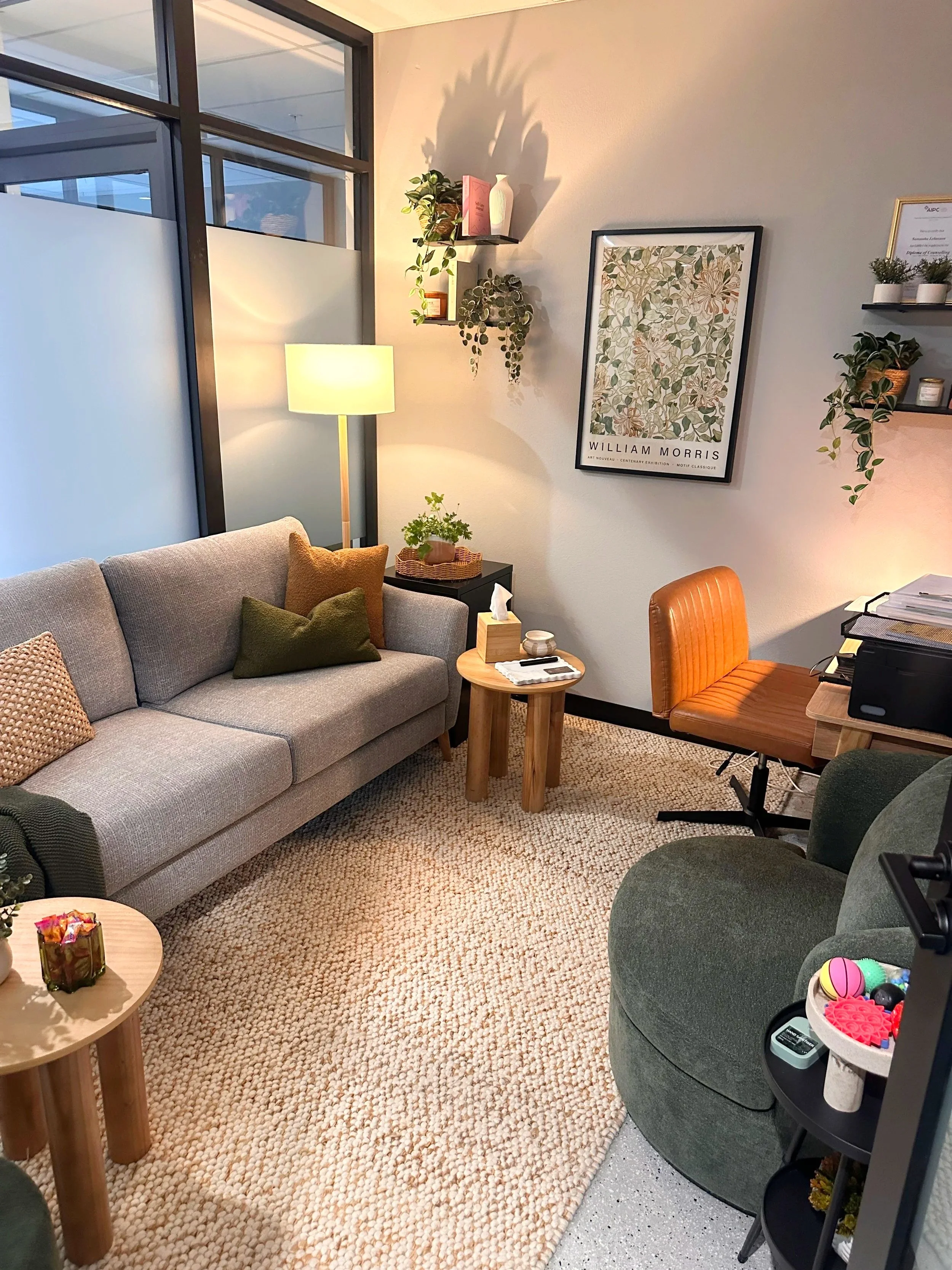 A cozy office space with a gray sofa adorned with green and beige pillows, a wooden side table, a lamp, and various plants on wall-mounted shelves, framed artwork, and a carpeted floor.