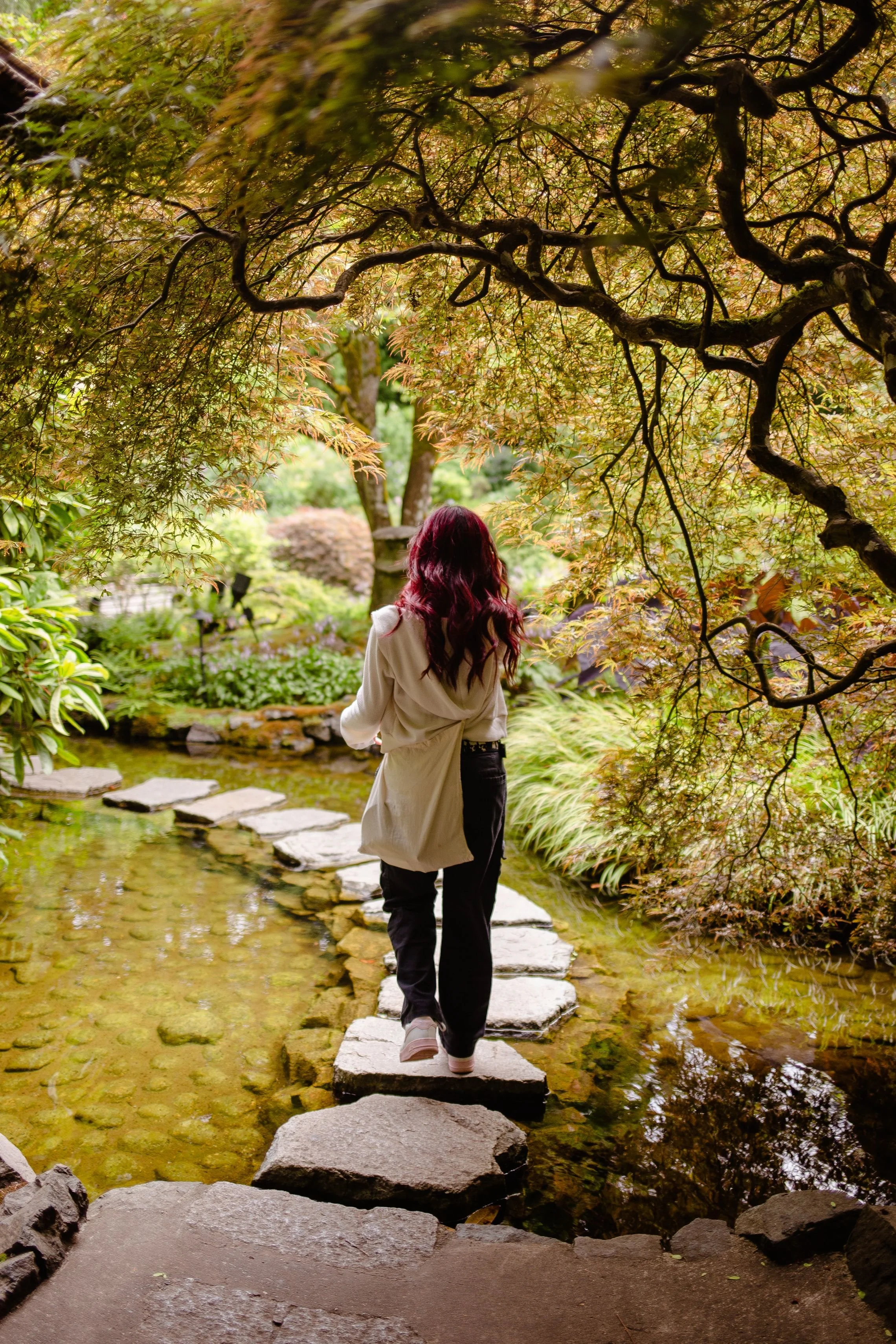 A person with long red hair, wearing a light-colored coat and black pants, walking barefoot across stepping stones in a small creek in a lush garden with green and orange foliage.