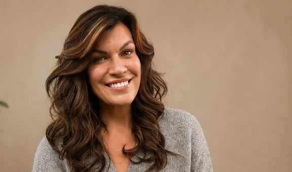 A woman with long, wavy brown hair smiling at the camera, wearing a gray top against a neutral background.