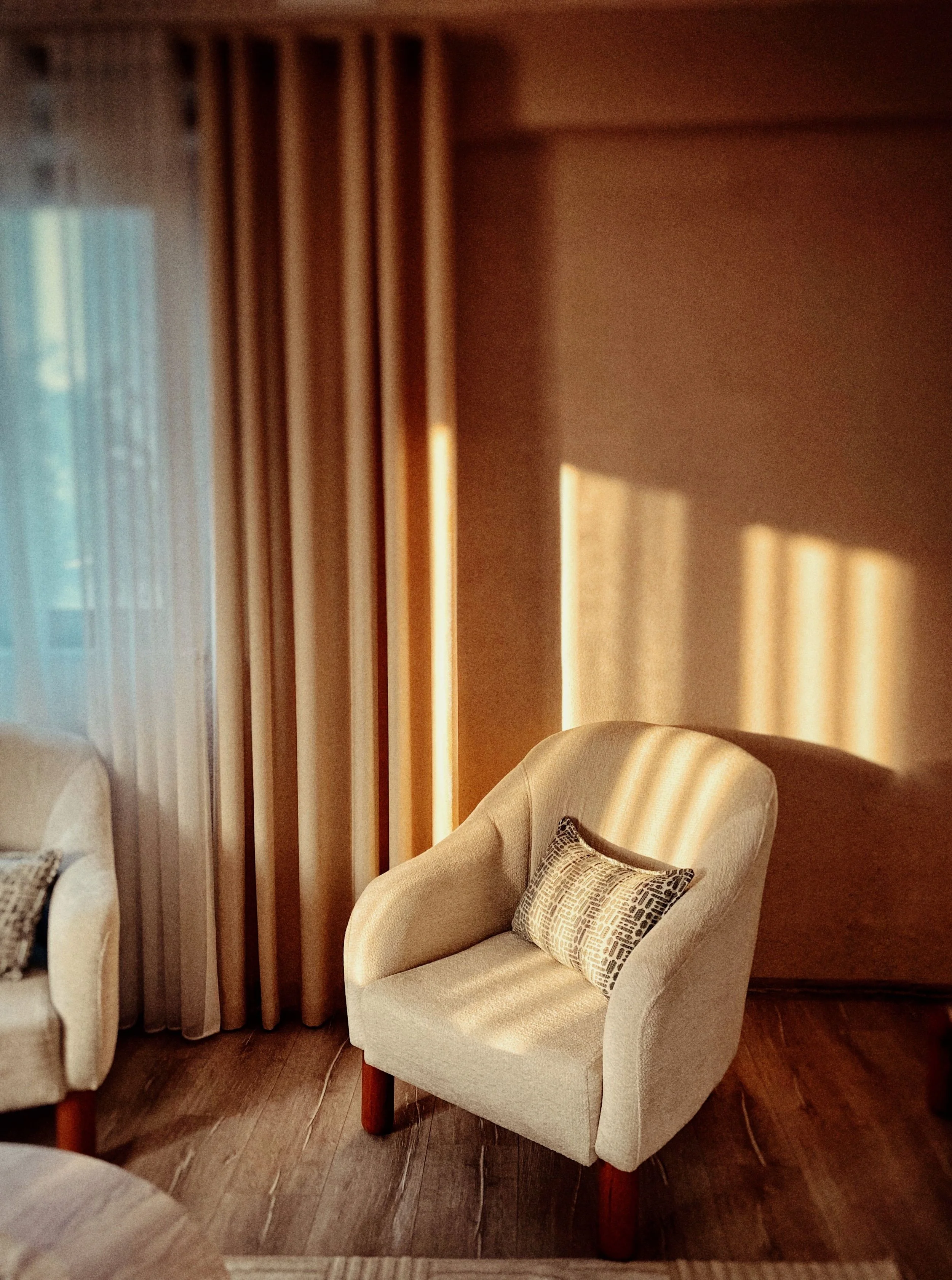 A cozy living room corner with an upholstered armchair, a decorative pillow, wooden flooring, beige curtains, and sunlight casting striped shadows across the wall.