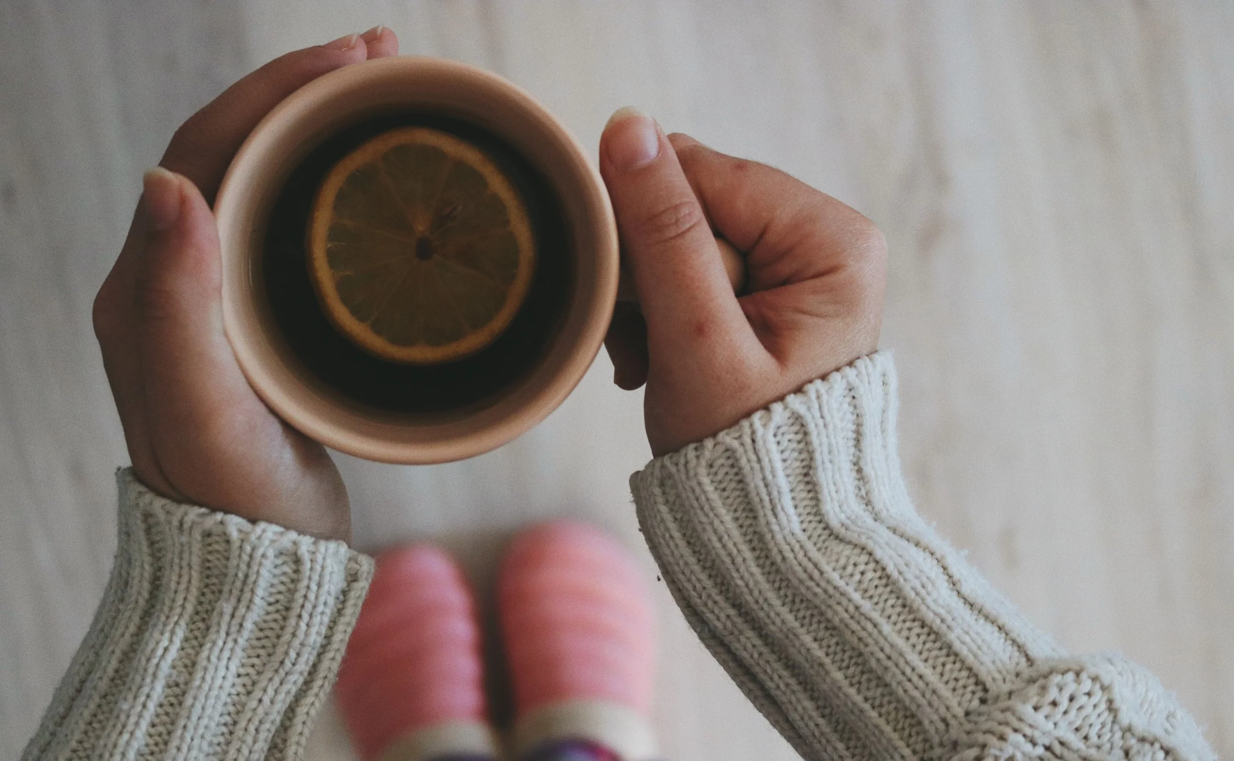 A person holding a cup containing a lemon slice, wearing a white knitted sweater.