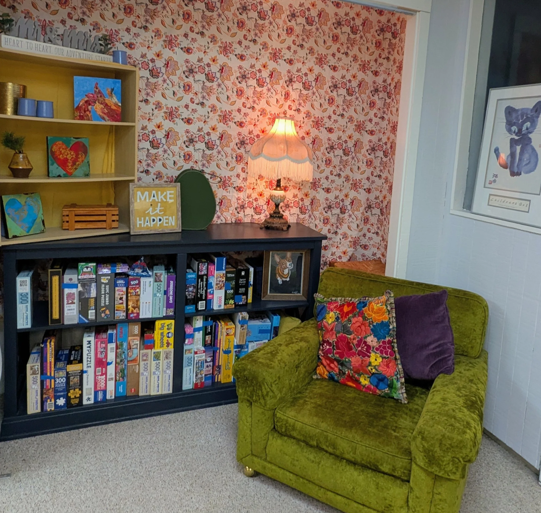 A room with a green velvet armchair with a floral patterned pillow, a yellow bookshelf with decorations and books, a vintage table lamp, and framed artwork on the wall. The background features floral wallpaper.