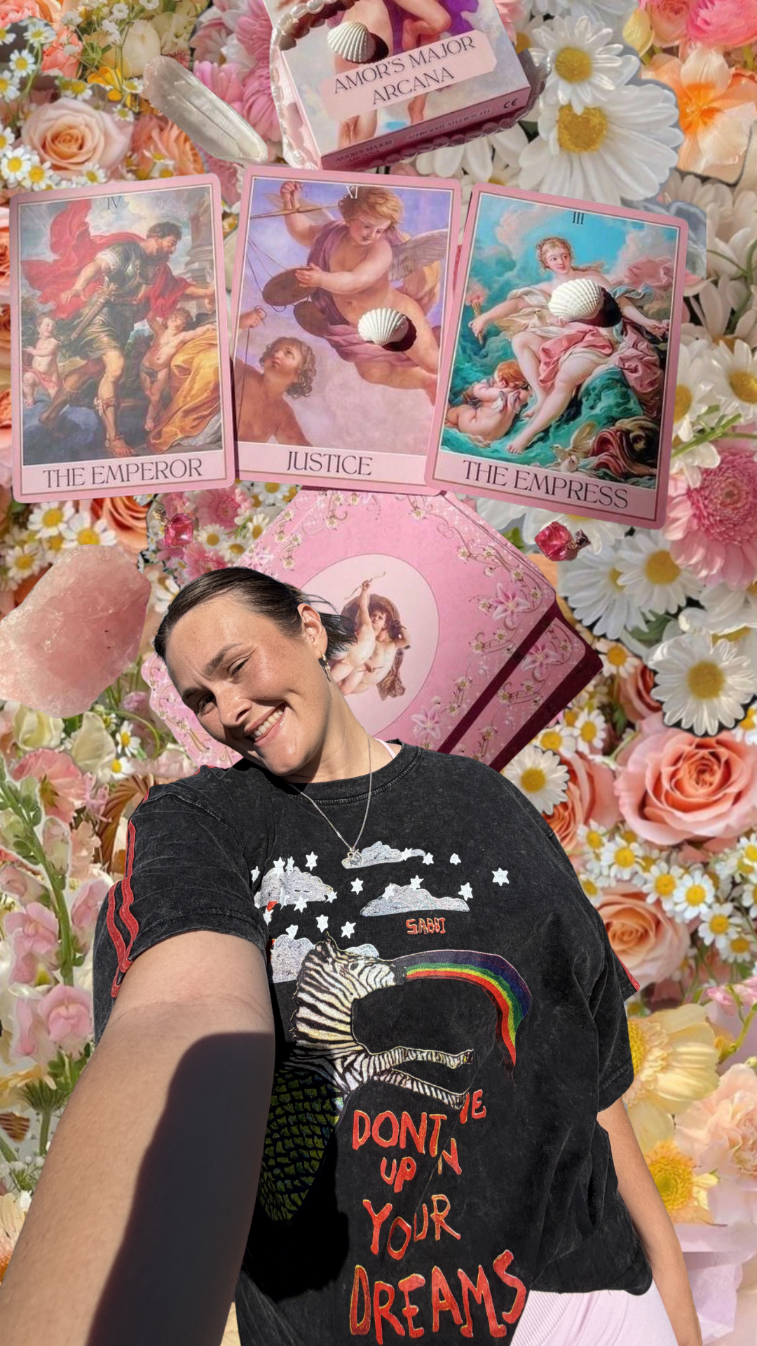 A woman taking a selfie on a floral background featuring cards depicting The Emperor, Justice, and The Empress tarot cards, along with flowers, a pink box labeled Amor's Major Arcana, and a person in the foreground wearing a black shirt with a rainbow and animal print design.